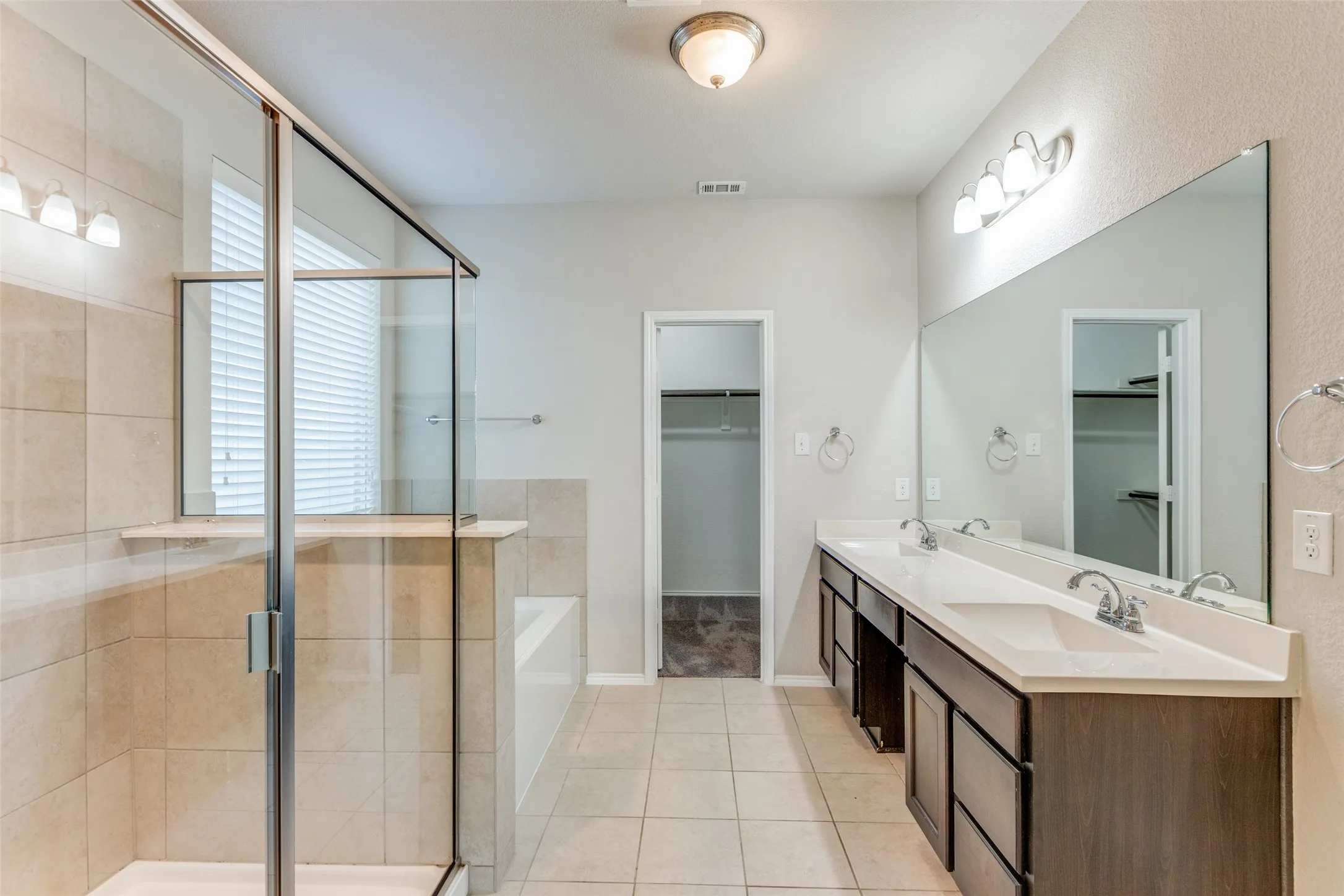 Primary bathroom featuring a spa-inspired layout with a frameless glass-enclosed walk-in shower, dual-sink vanity with wood-finish cabinetry and white countertops, large illuminated mirror, and ceramic tile flooring throughout