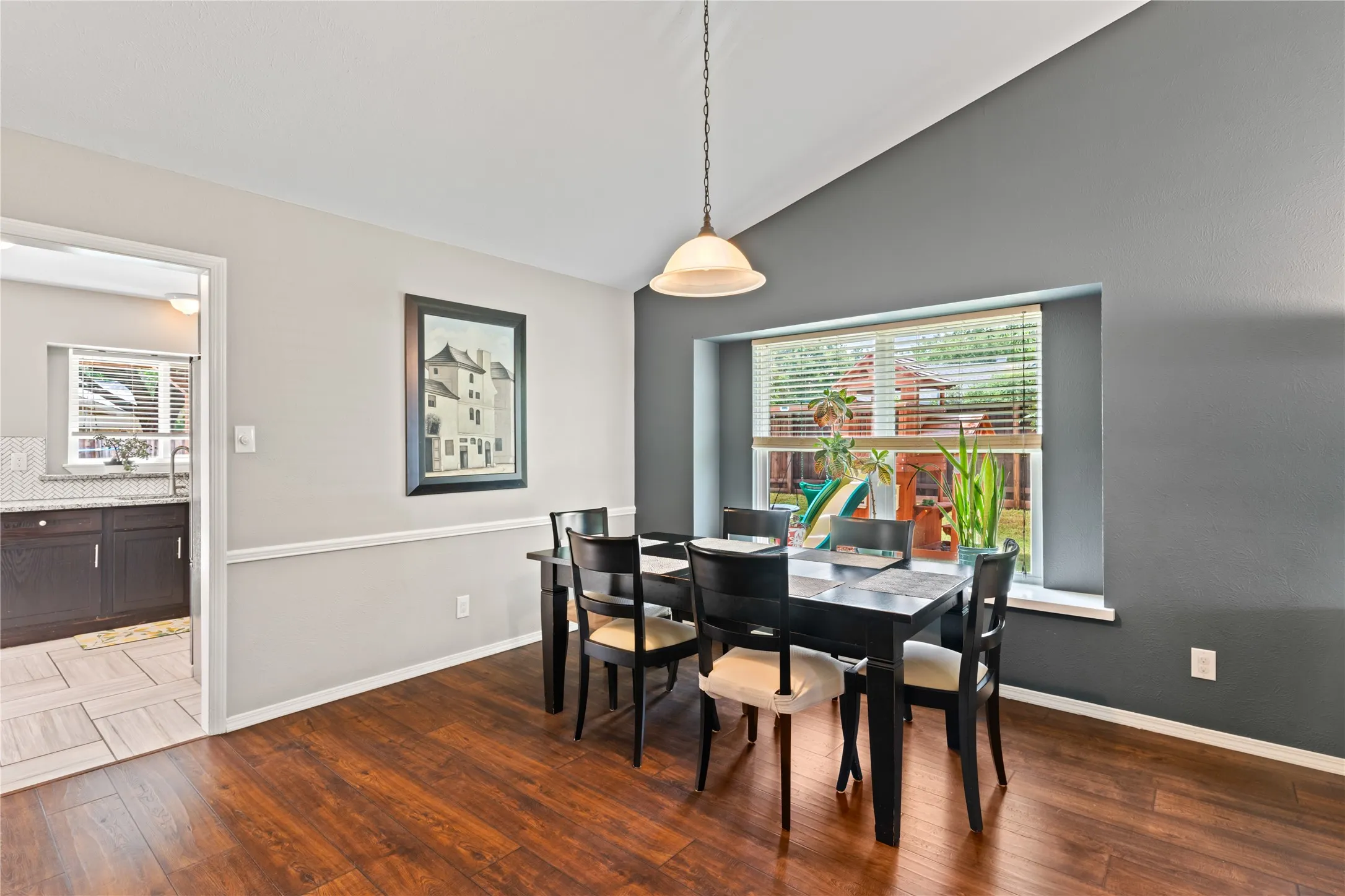 Vaulted ceiling with pendant lighting, wood-finish flooring, two-tone wall treatment, and a large window with blinds