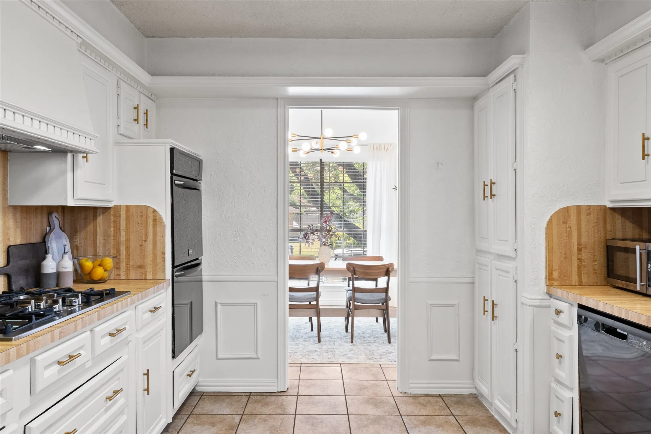 White cabinetry with gold-finish hardware, wood-finish countertops, tiled flooring, wainscoting details, and a built-in oven