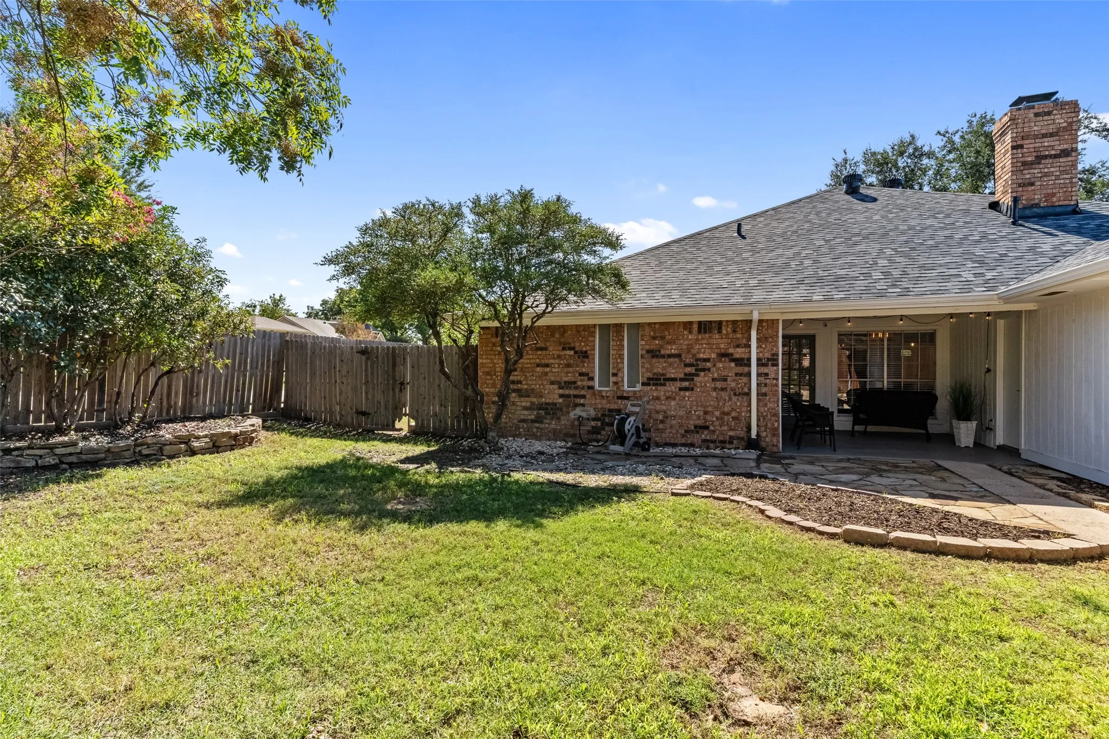 Fenced backyard with a covered patio, brick chimney, and mature landscaping