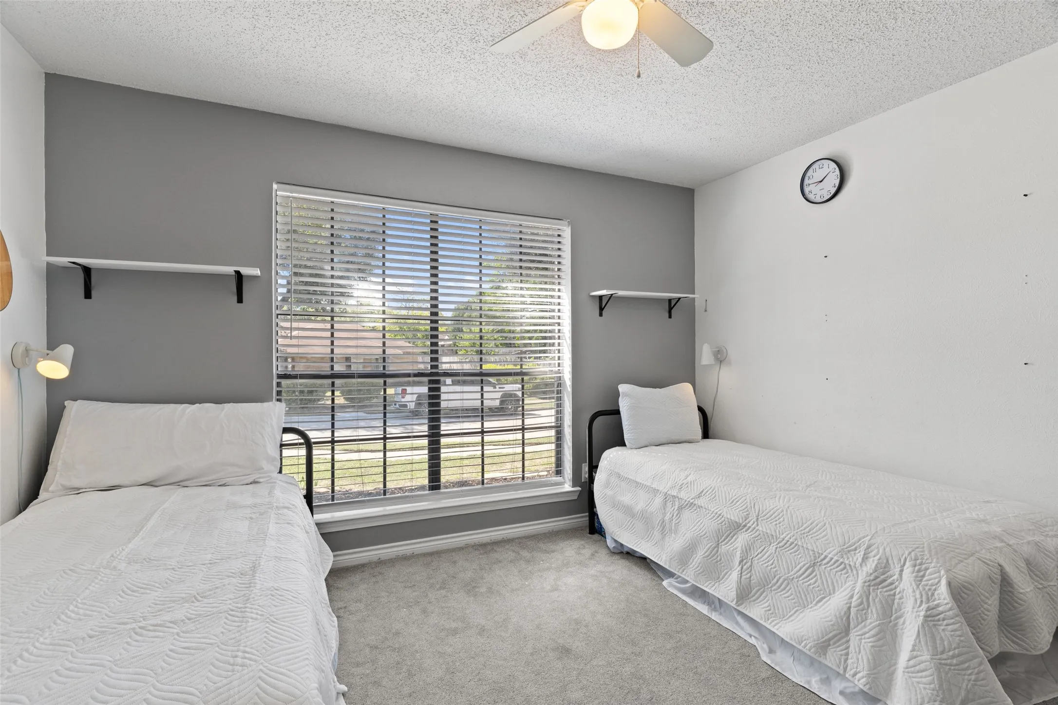 Carpeted flooring, window with blinds, ceiling fan with integrated lighting, wall-mounted shelving units, and sconce lighting