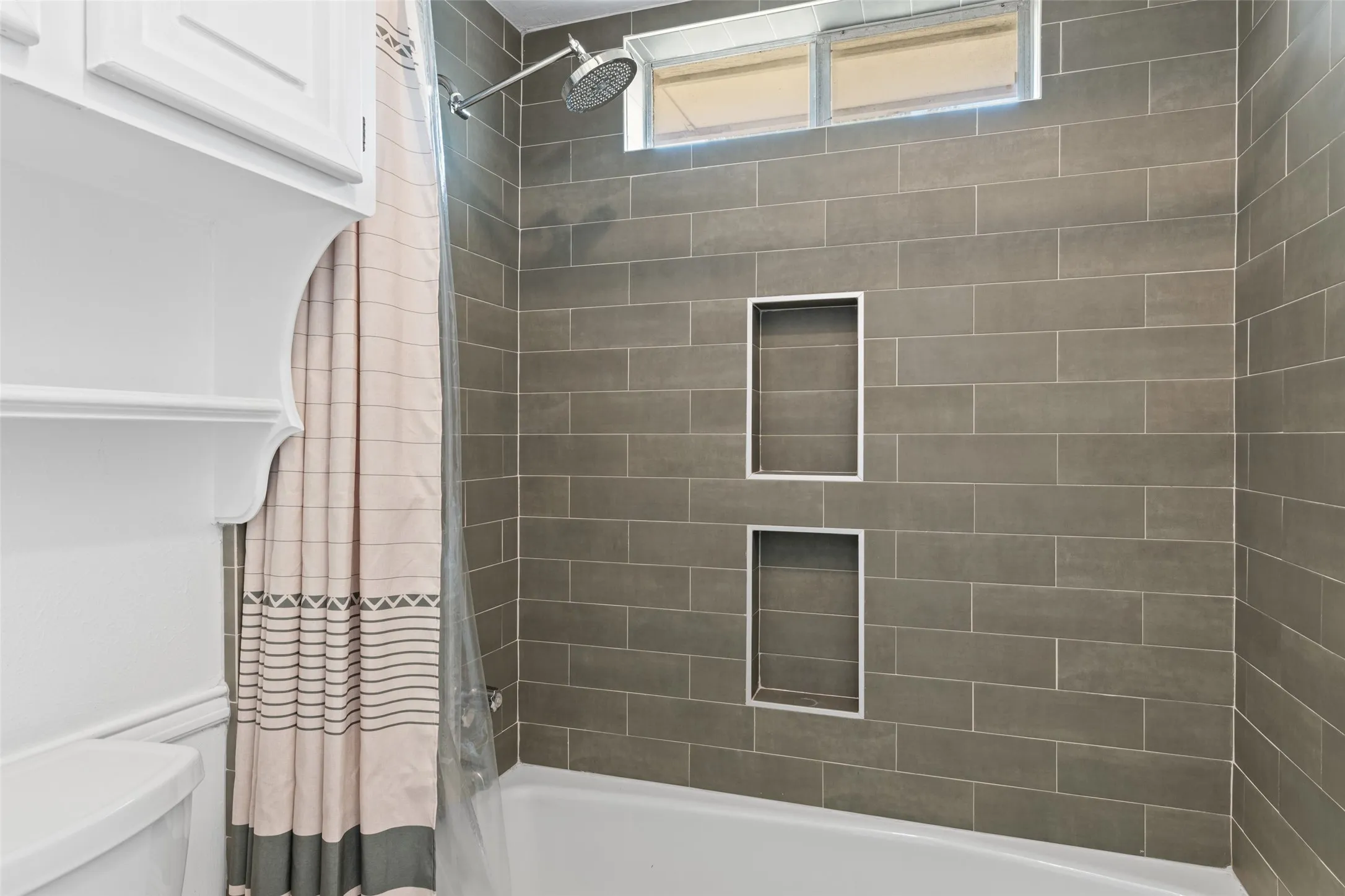 Bathtub with subway tile surround, rain shower head, dual recessed shower niches, and an overhead transom window