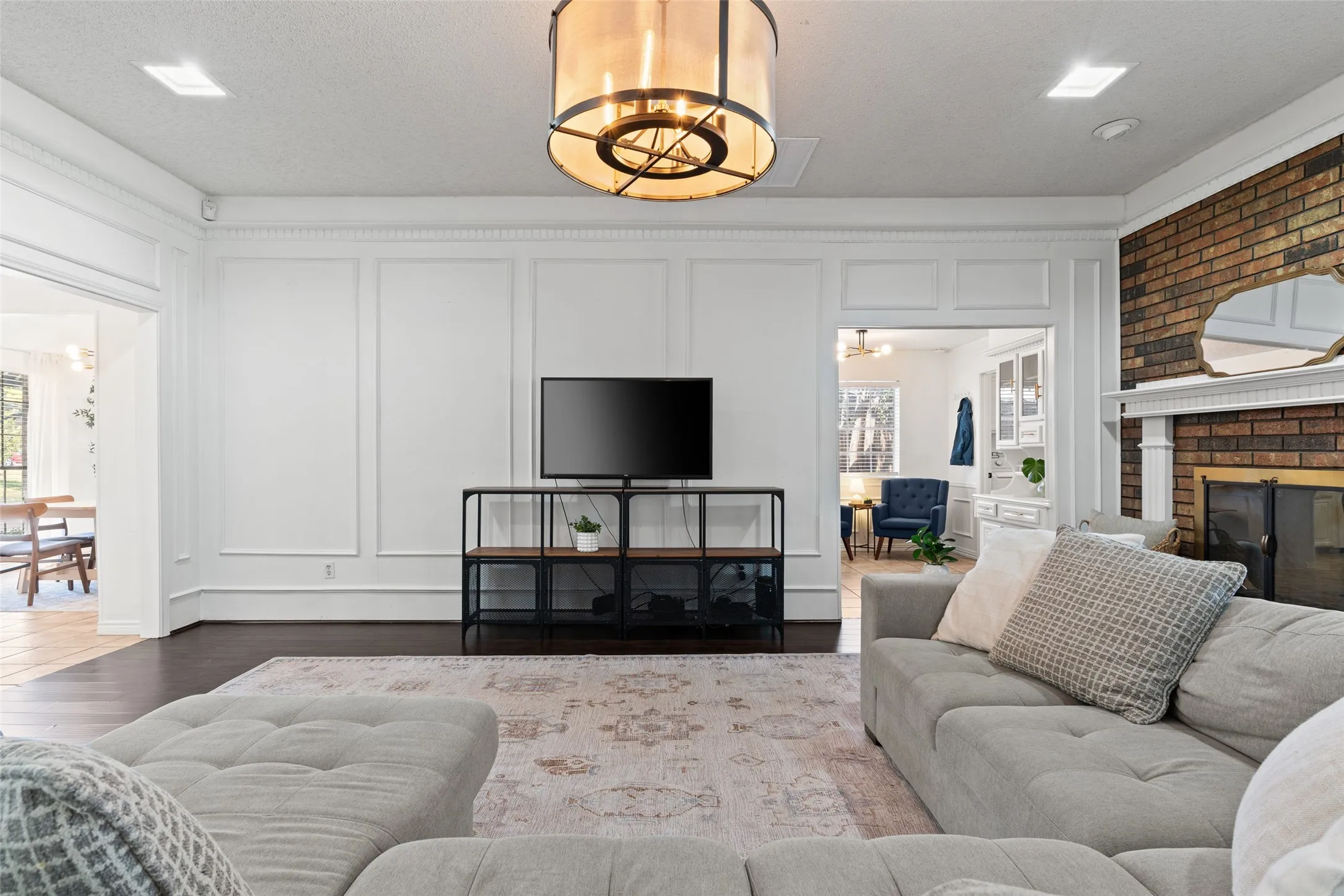 Hardwood flooring, decorative wall paneling, recessed lighting, brick fireplace with a white mantel, and a prominent ceiling light fixture