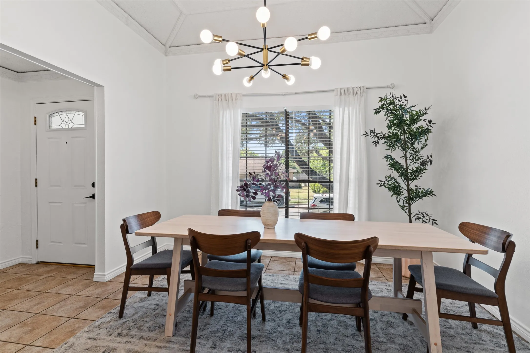 Vaulted ceiling with decorative beams, modern sputnik chandelier, large window with blinds and sheer curtains, paneled entry door with decorative glass insert, and tile flooring
