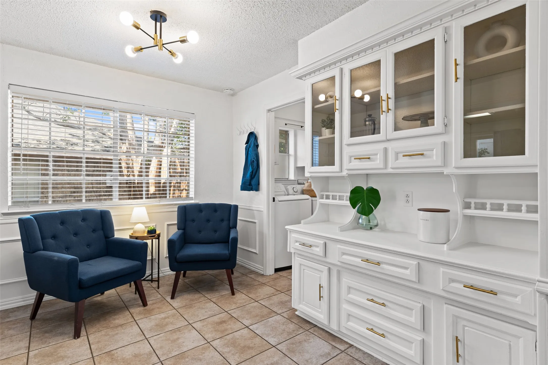 White built-in cabinetry with glass-front upper doors and gold-finish hardware, tile flooring, and a large window with blinds