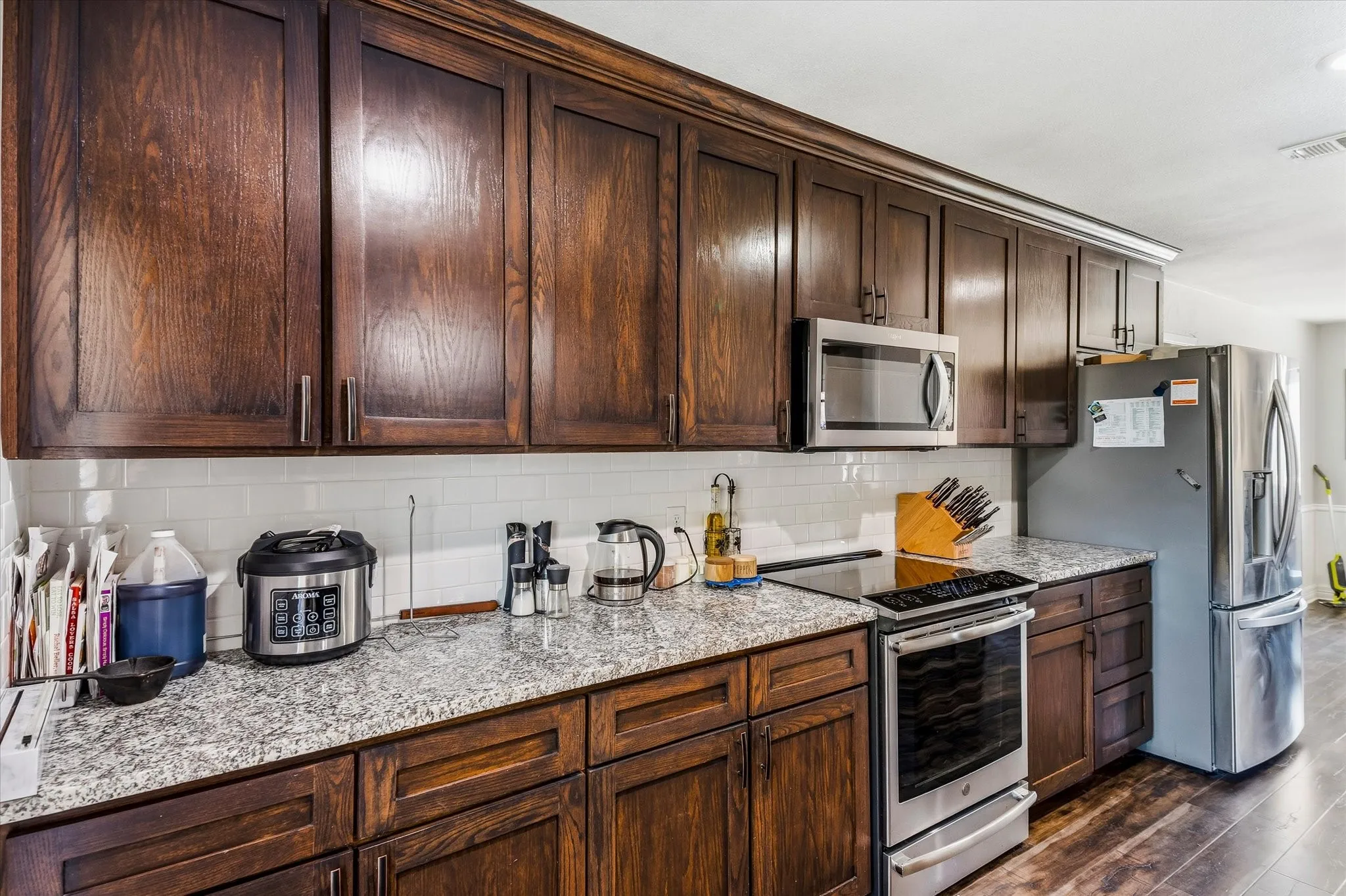 Dark wood cabinetry, speckled countertops, stainless steel appliances, white subway tile backsplash, wood-finish flooring