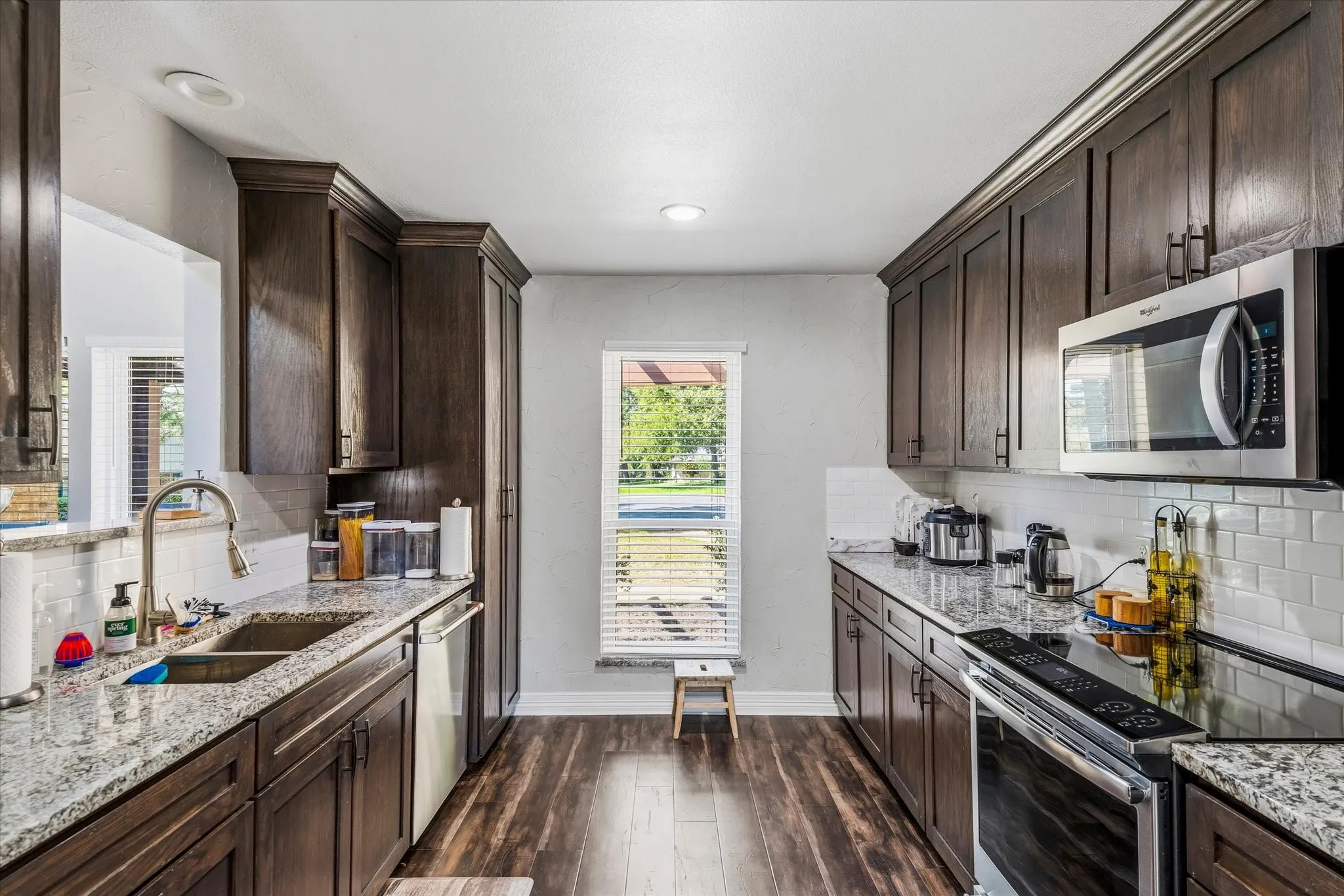Galley kitchen featuring wood-finish flooring, dark wood cabinetry, light-toned granite countertops, and a subway tile backsplash