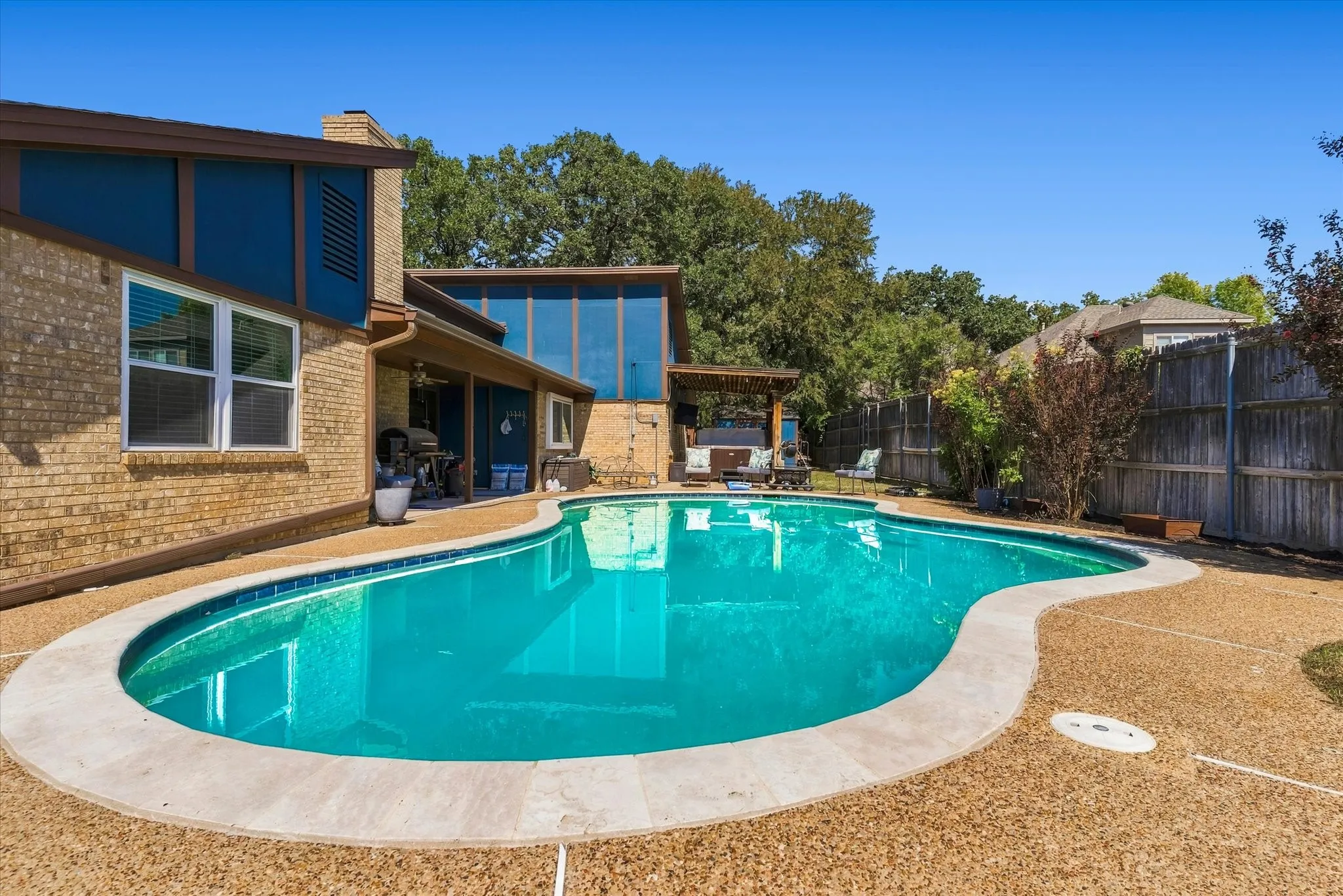 Curved swimming pool with stone coping, textured aggregate deck, private backyard fencing, and an integrated covered patio structure