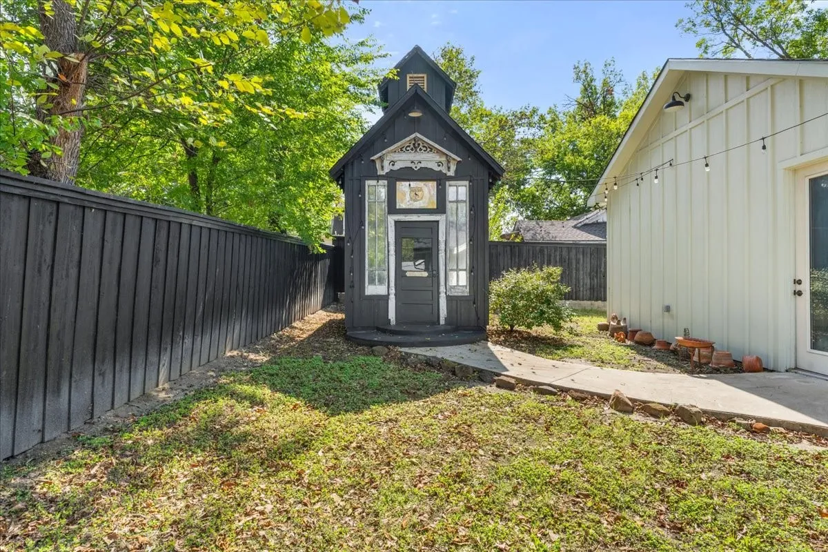 Black vertical board fence, concrete pathway, and a detached structure with a white-trimmed door and decorative facade