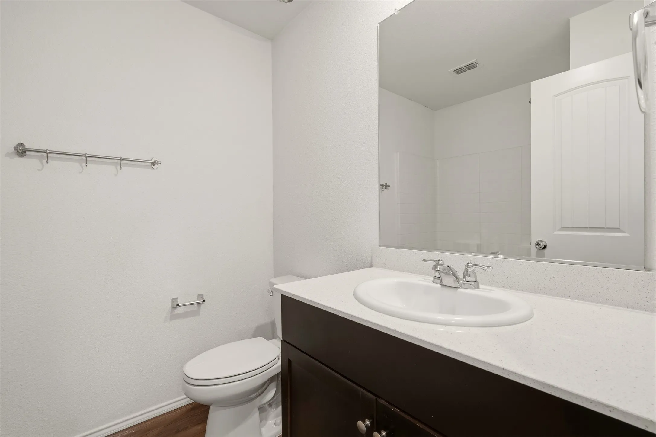 Wood-finish flooring, dark wood vanity with white countertop, integrated sink with chrome faucet, large wall-mounted mirror, and white toilet