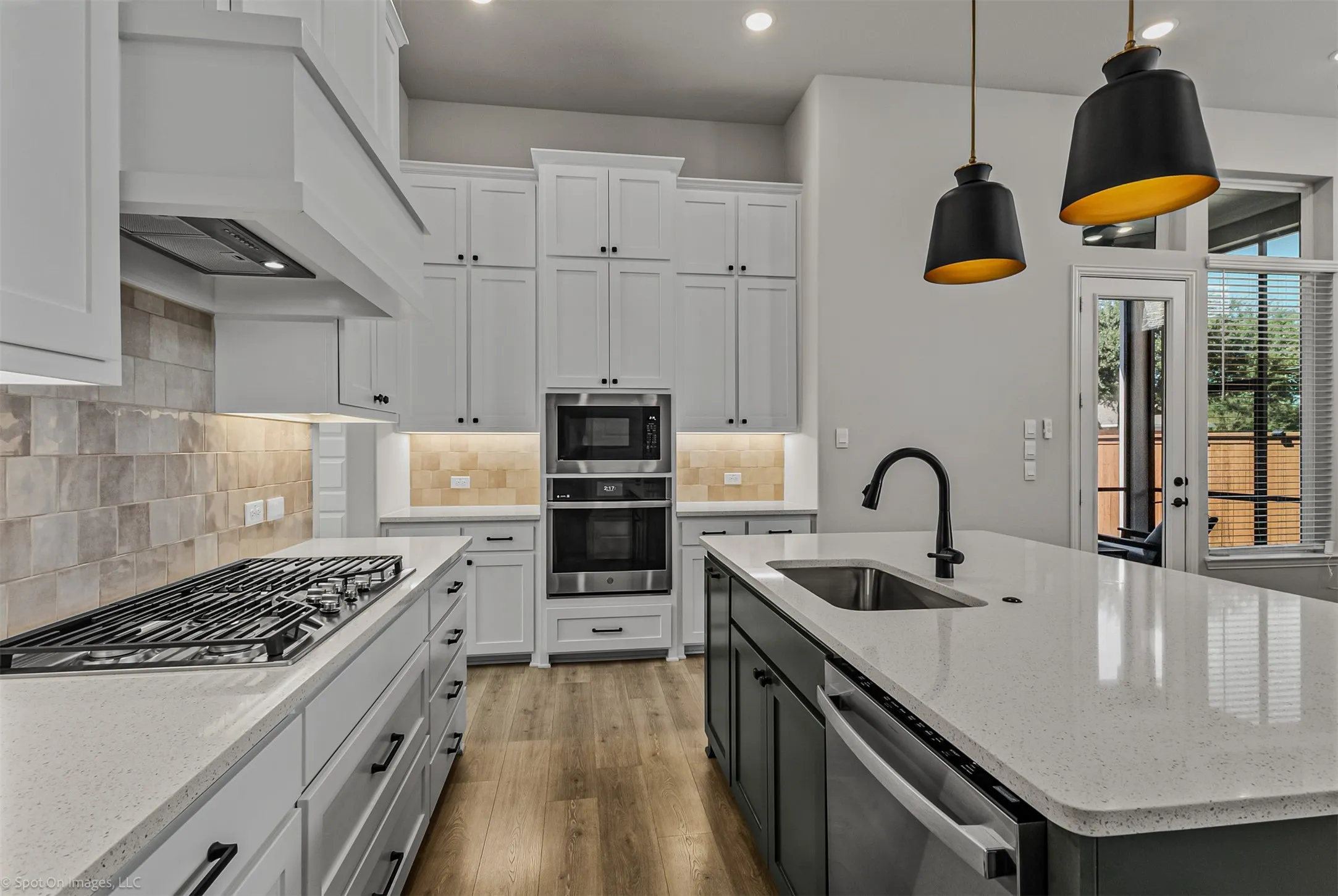 White shaker cabinetry, light-toned tiled backsplash, stainless steel appliances, large kitchen island with integrated sink, and wood-finish flooring