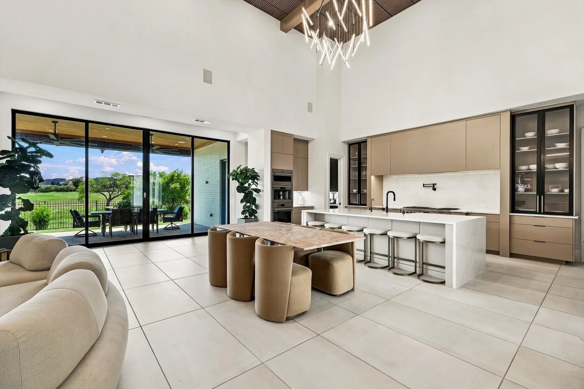 Expansive main living area featuring high vaulted ceilings, large format tile flooring, a modern kitchen with a waterfall island, and floor-to-ceiling glass panel doors opening to an outdoor patio