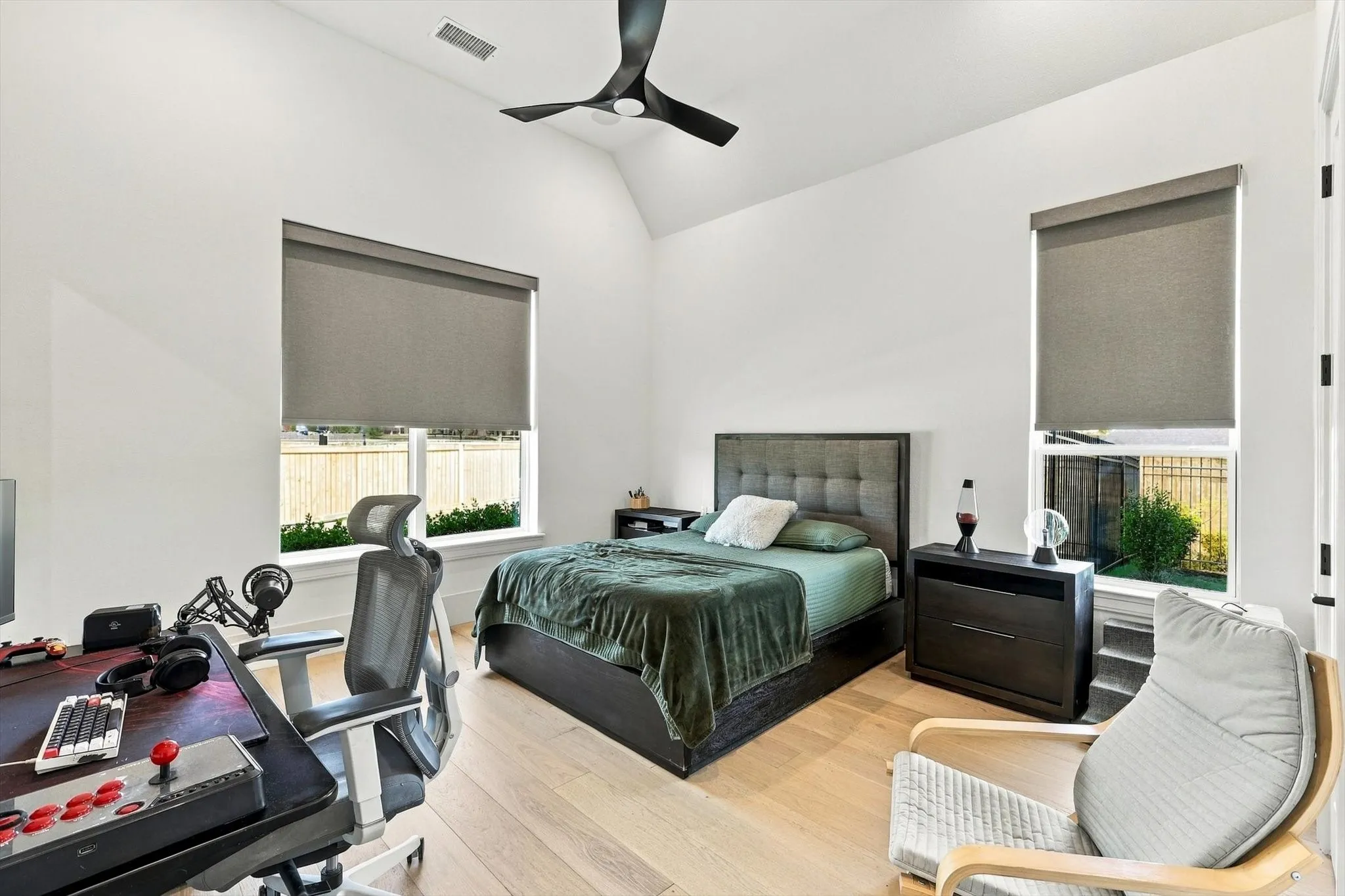 Vaulted ceiling with a contemporary ceiling fan, light wood-finish flooring, two large windows with roller shades, and a built-in bed frame with a tufted headboard