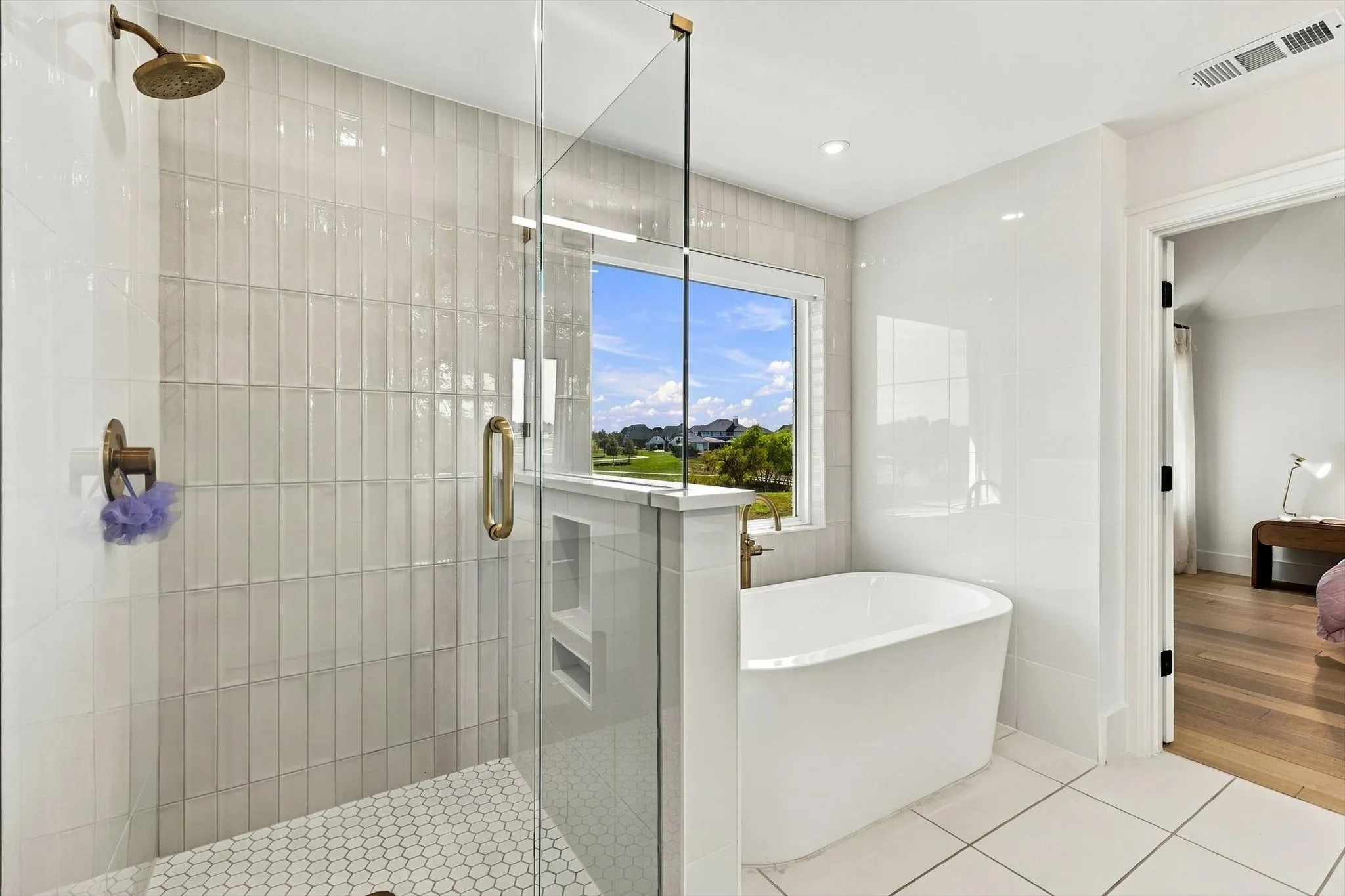 Frameless glass shower with brass fixtures, a freestanding soaking tub, and a window providing natural light