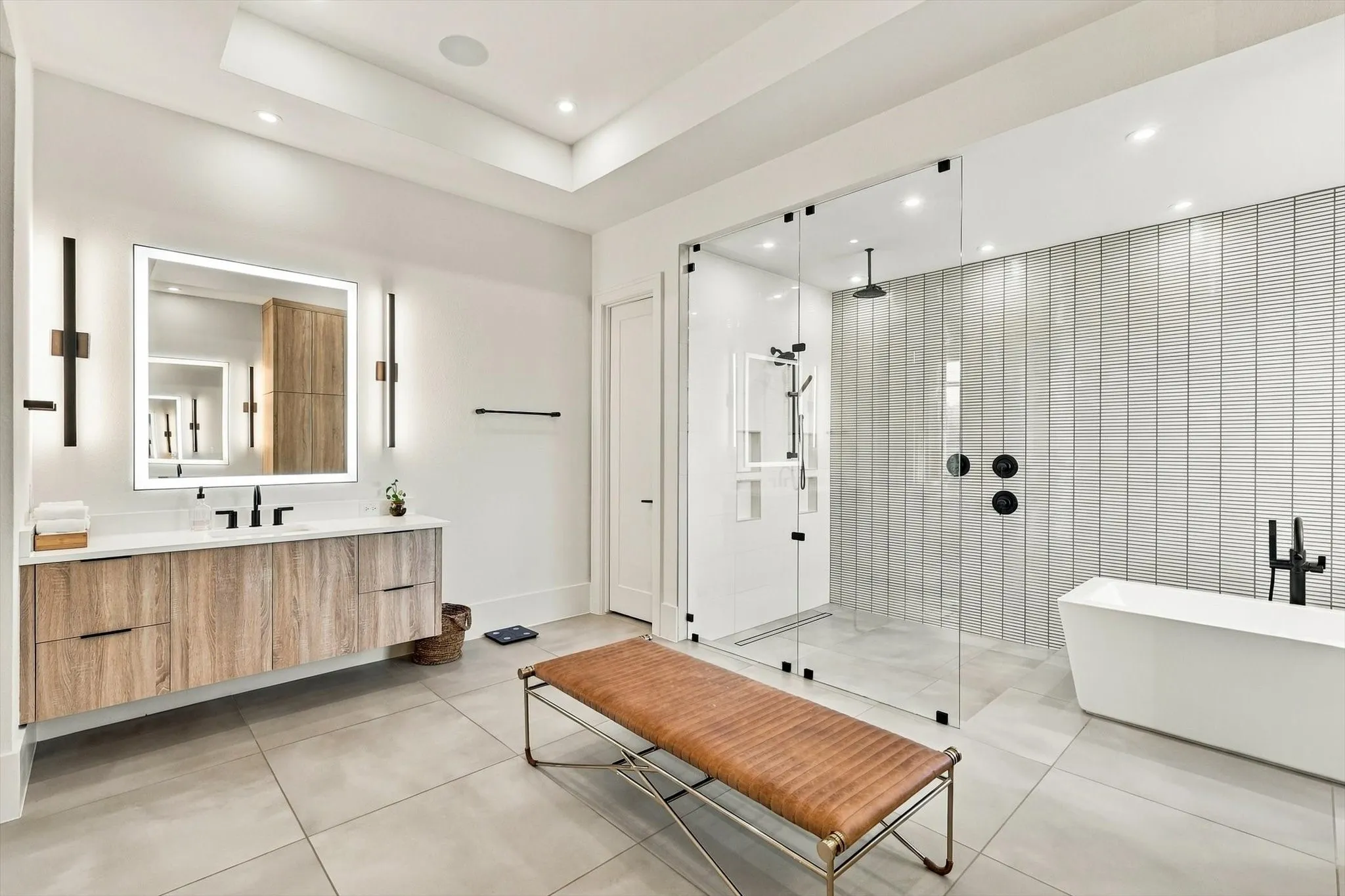 Floating wood-finish vanity with an illuminated mirror, glass-enclosed shower, freestanding bathtub, and large format floor tiling