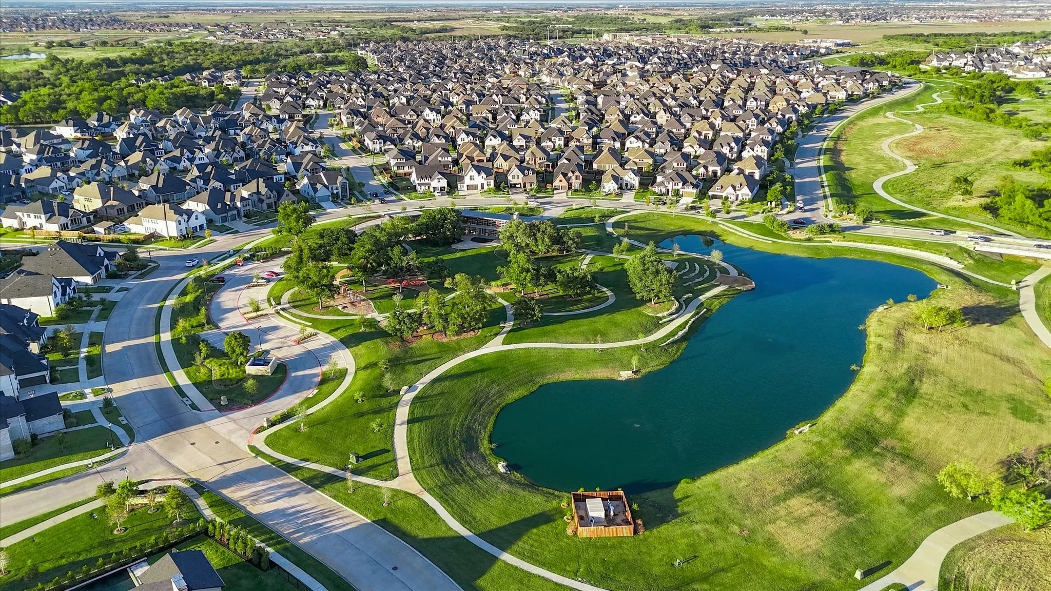 Expansive community featuring residential properties, a large central pond, numerous paved walking paths, and extensive green spaces with mature trees
