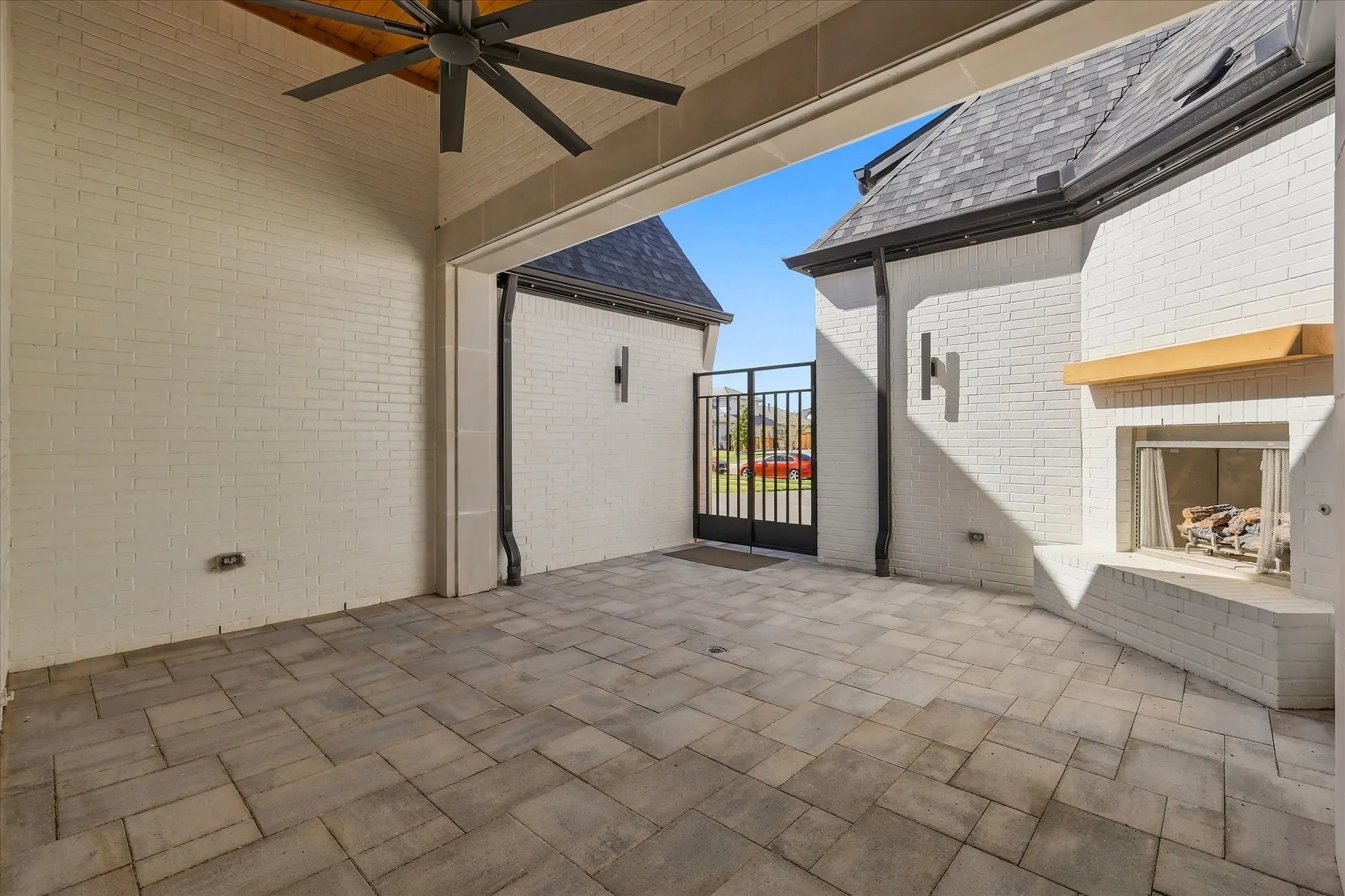 Covered outdoor patio featuring a ceiling fan, brick fireplace with wood mantle, paver flooring, and a metal gate