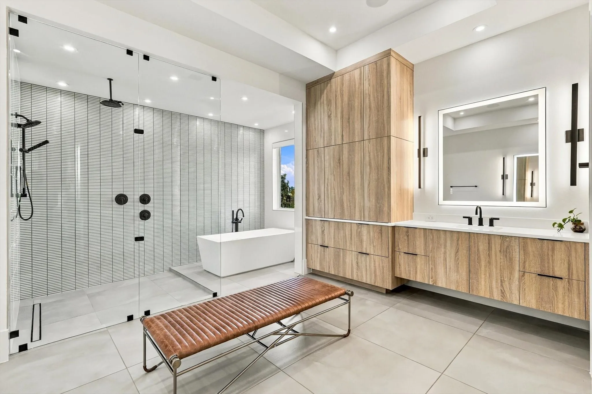 Spacious bathroom featuring a glass-enclosed shower with multiple showerheads, a freestanding soaking tub, wood-finish cabinetry, a large vanity with an integrated mirror, and light-toned tile flooring