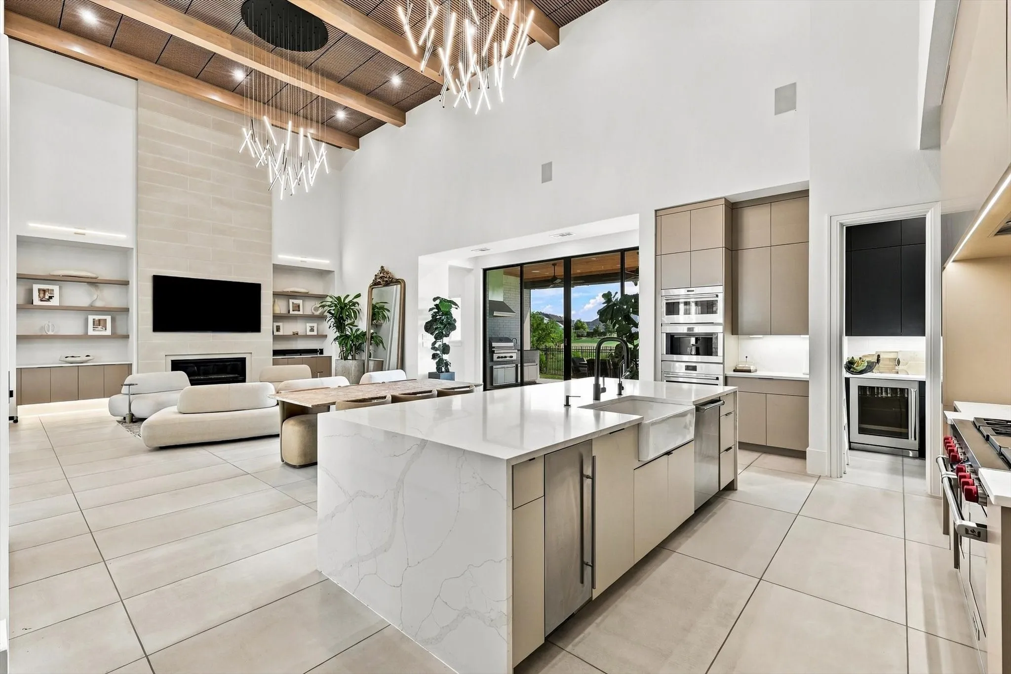 Two-story ceiling with exposed wood beams and contemporary chandeliers, stone fireplace with built-in shelving, expansive kitchen island with a farmhouse sink, integrated stainless steel appliances, and large format tile flooring
