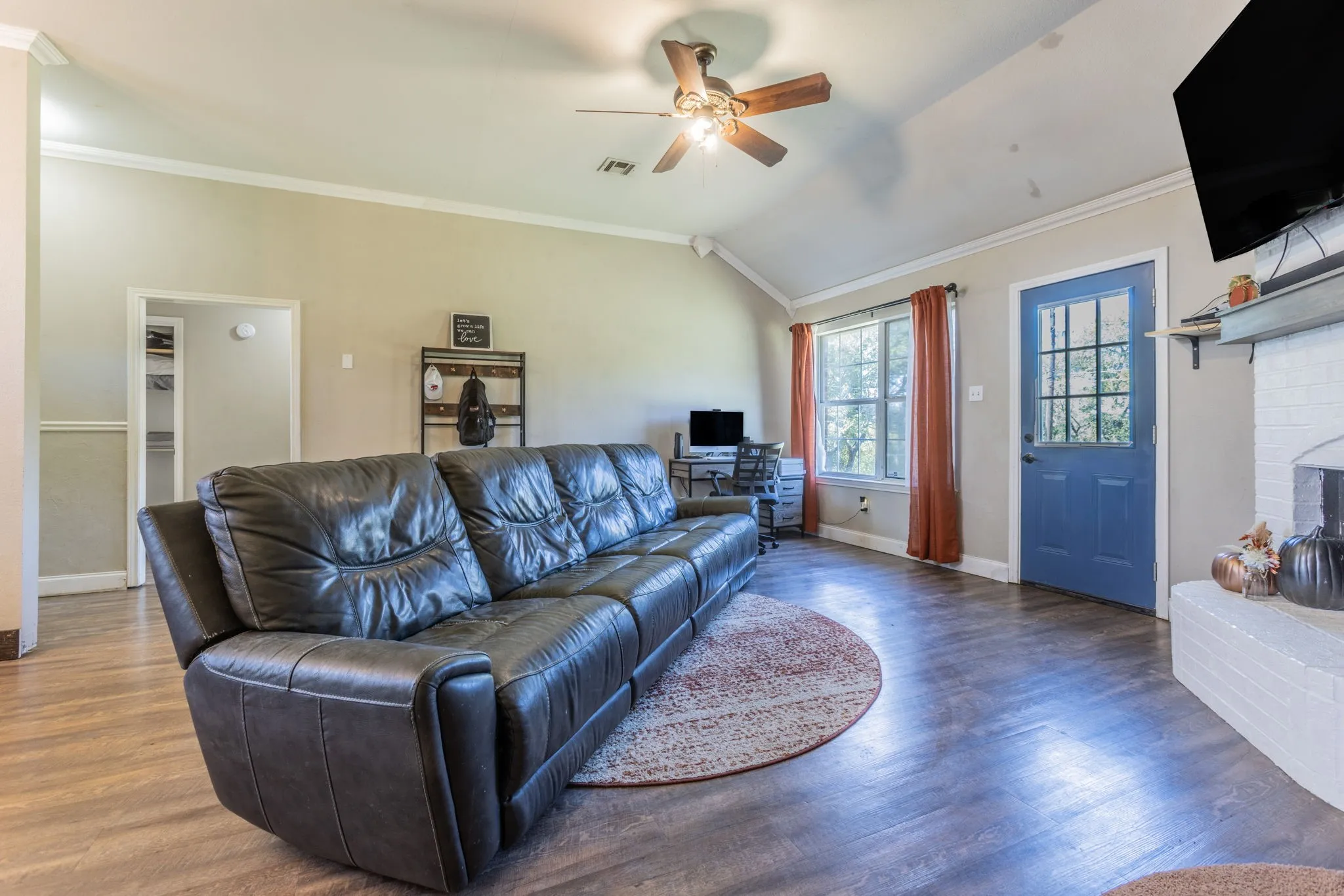Vaulted ceiling, wood-finish flooring, white brick fireplace, ceiling fan, and a blue paneled door with glass inserts