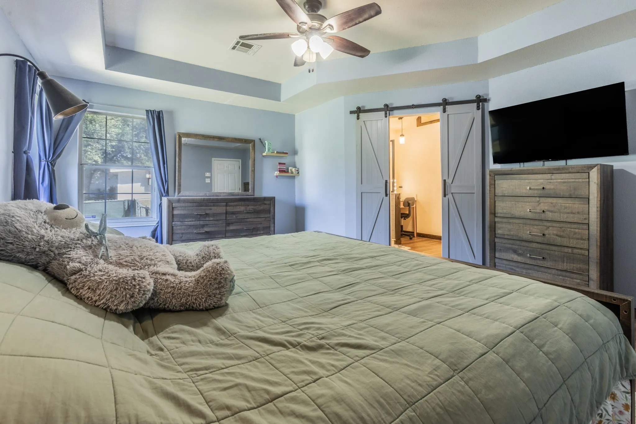 Tray ceiling with a ceiling fan, window with exterior views, barn door entry, and wood-finish flooring visible beyond the barn doors