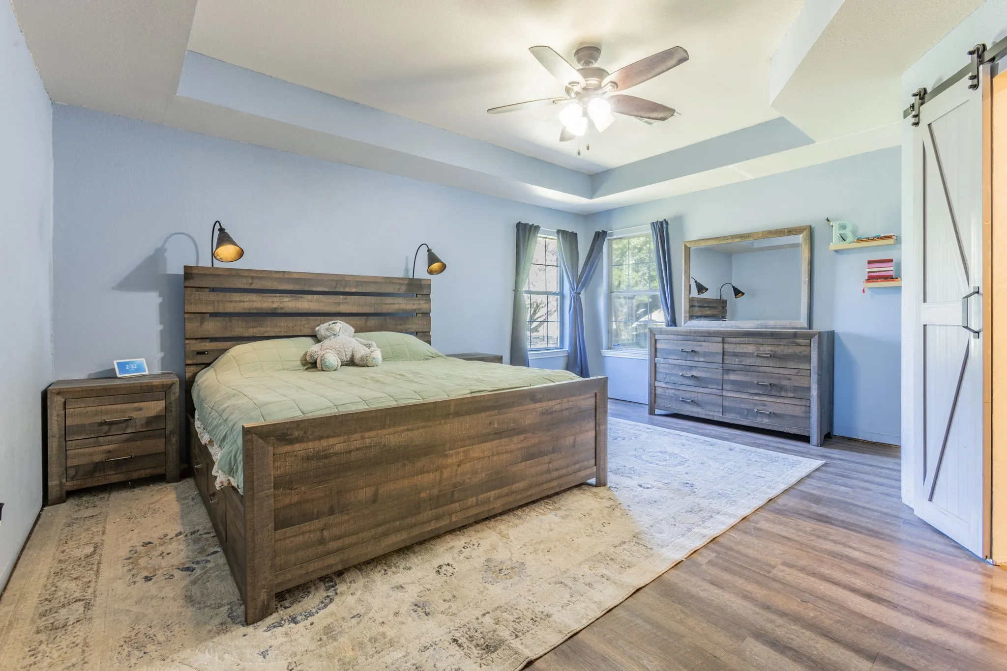 Tray ceiling with a ceiling fan, wood-finish flooring, two windows, and a barn door