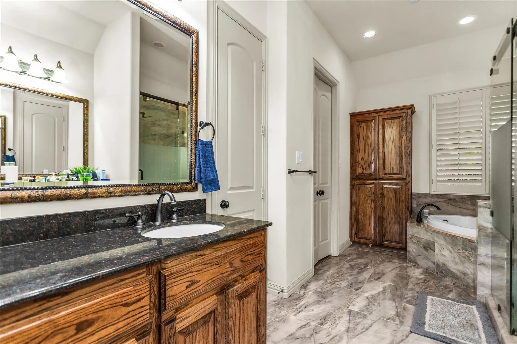 Vanity with a dark countertop and undermount sink, framed mirror, built-in wooden cabinet, soaking tub with tile surround, and plantation shutters