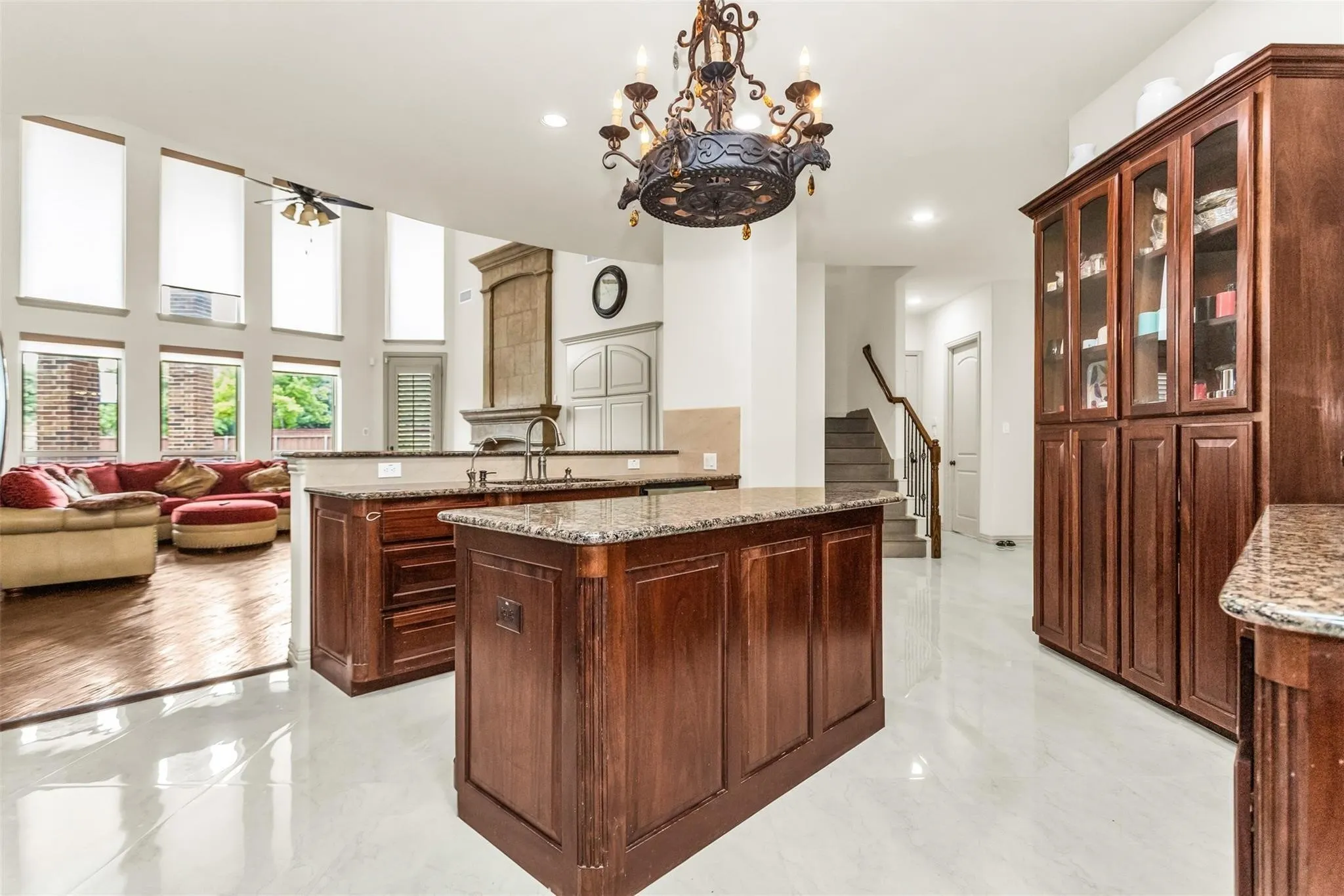Central kitchen islands with granite countertops, rich wood cabinetry with glass-front display, white tile flooring, and high ceilings with upper windows