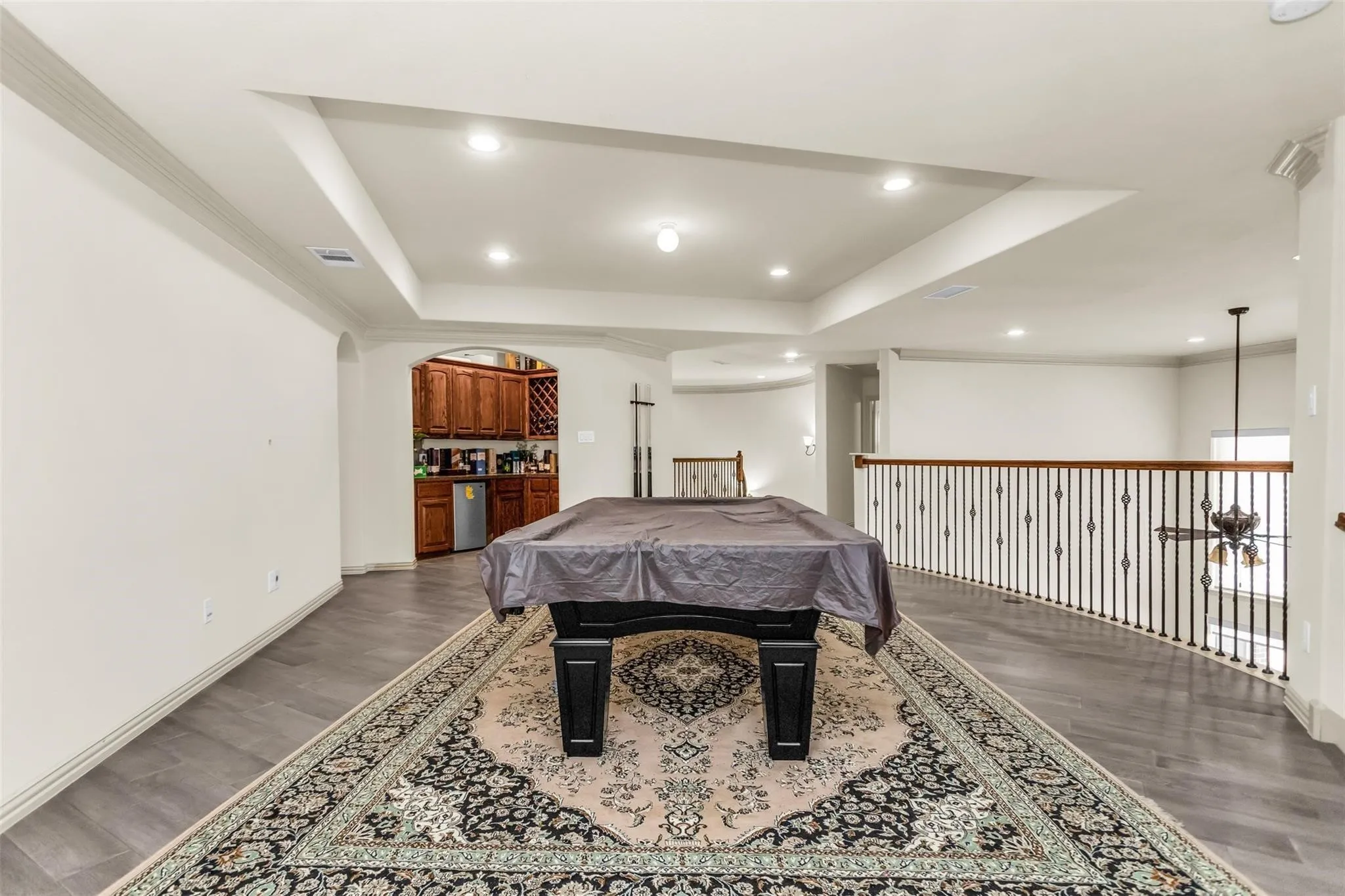 Tray ceiling with recessed lighting, wood-finish flooring, built-in wet bar with wood cabinetry, and wrought iron railing
