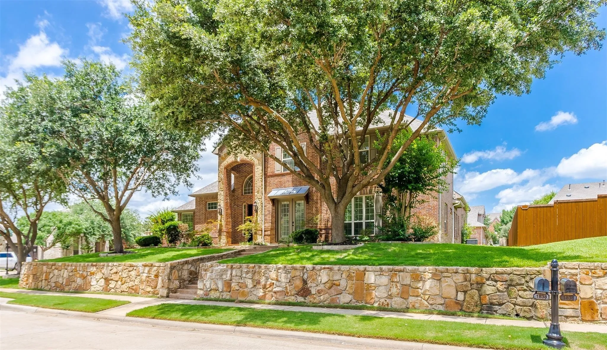 Traditional brick residence featuring an arched portico entry, mature trees, manicured lawn, and a stone retaining wall with integrated steps