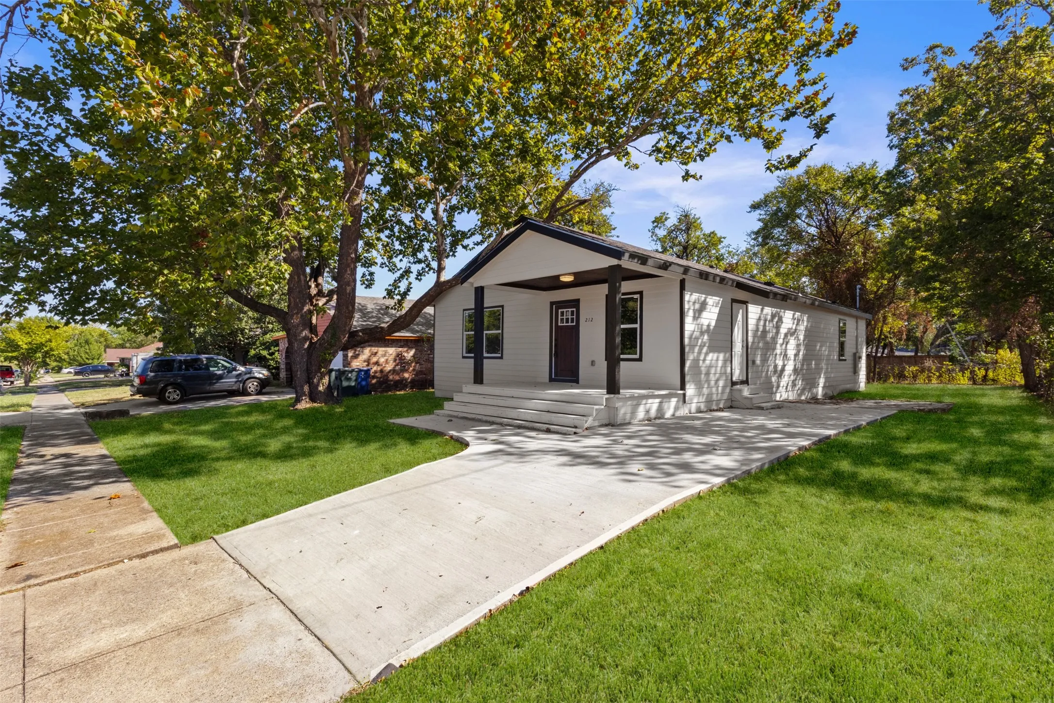 White siding exterior with dark trim accents, covered front porch, concrete driveway, and a mature tree