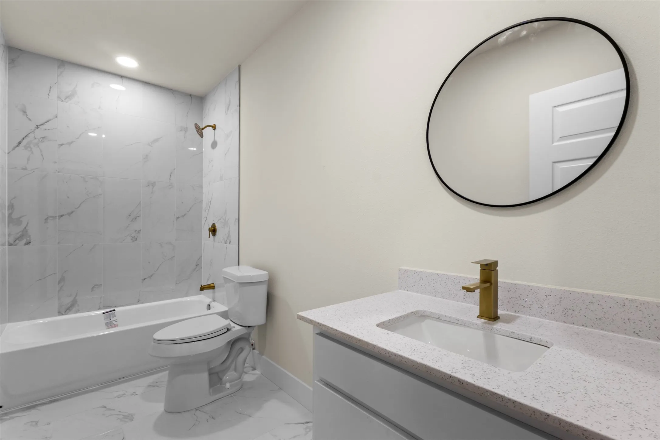Marble-look tile flooring, white bathtub with a bronze shower fixture, white vanity with a bronze faucet, white countertop with integrated sink, and a circular mirror with a black frame