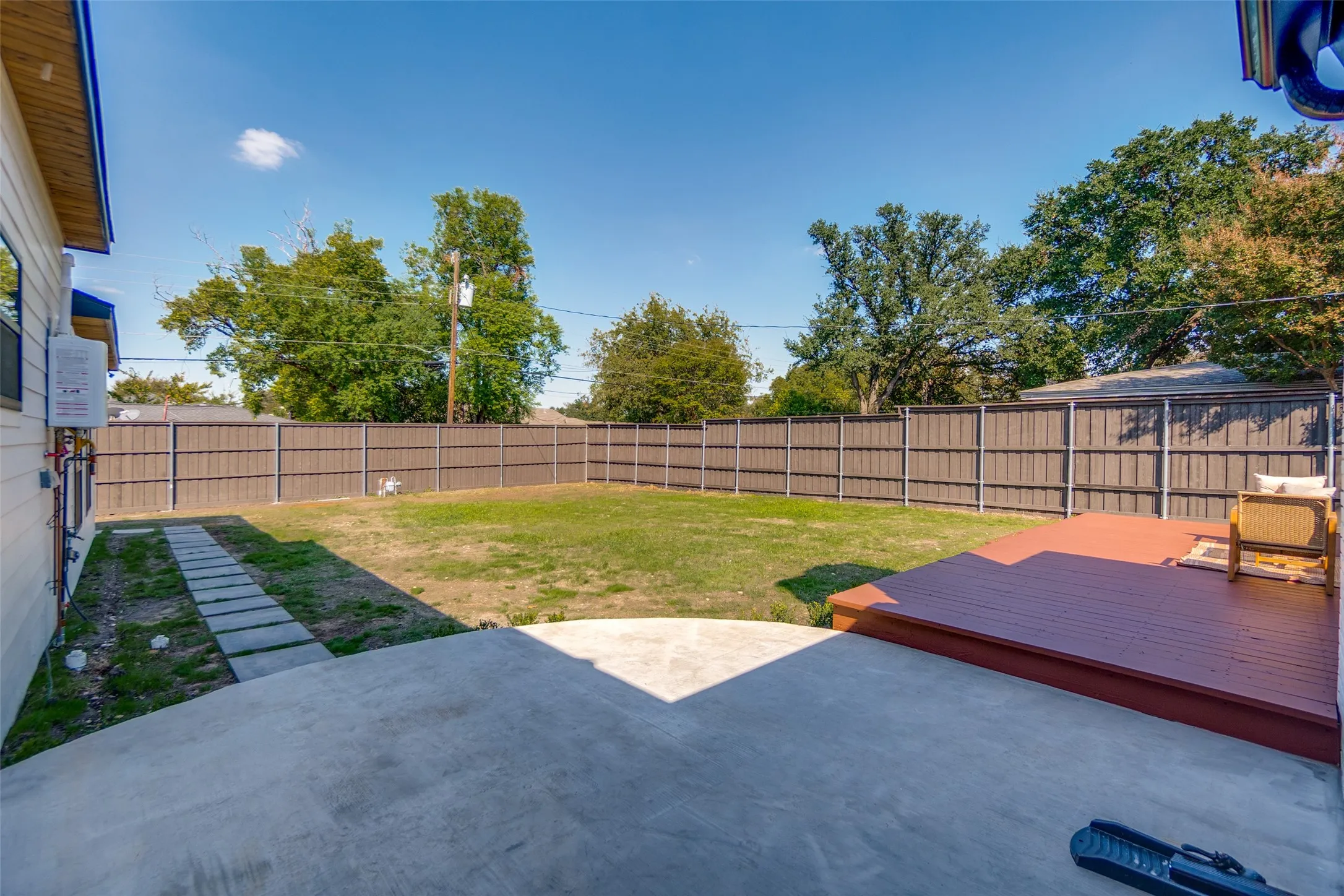 Expansive backyard featuring a concrete patio, a raised wood deck, a full privacy fence, a manicured lawn, and mature trees