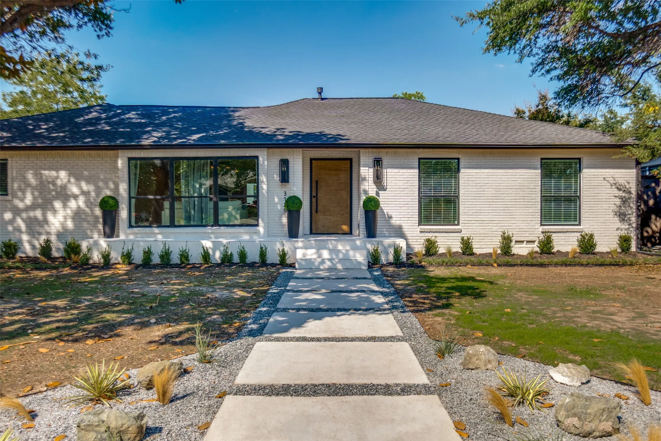 White brick exterior, black-framed windows, wood-finish entry door, concrete paver walkway, and a landscaped front yard