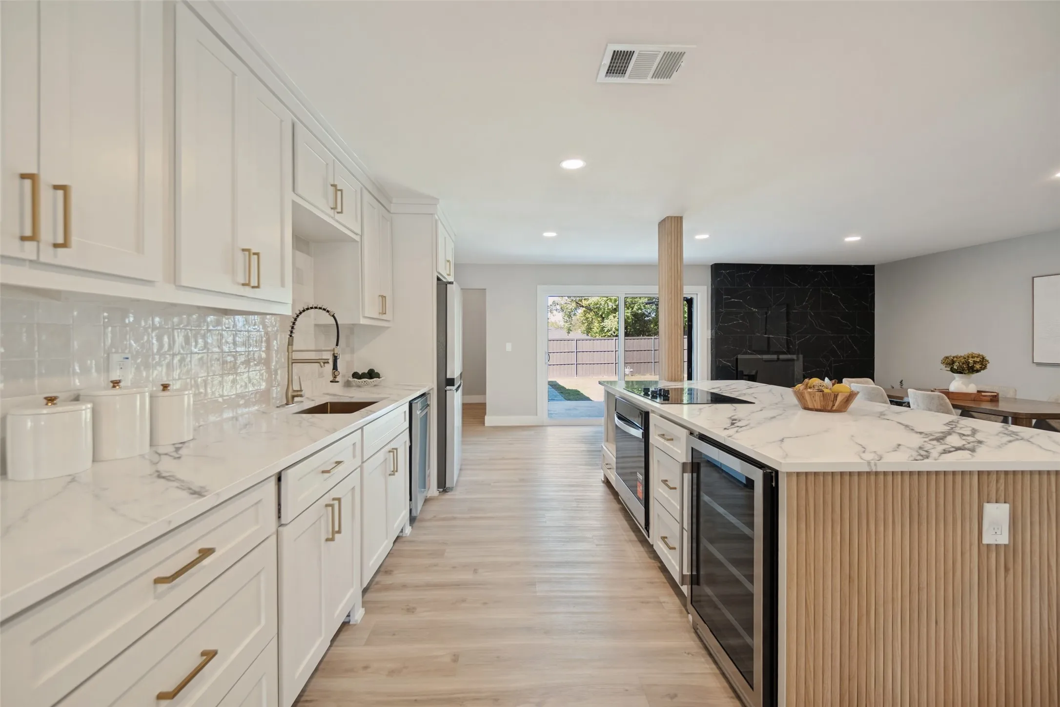 White shaker cabinetry with gold-finish hardware, white marbled countertops, light wood-finish flooring, a black marbled accent wall, and recessed lighting
