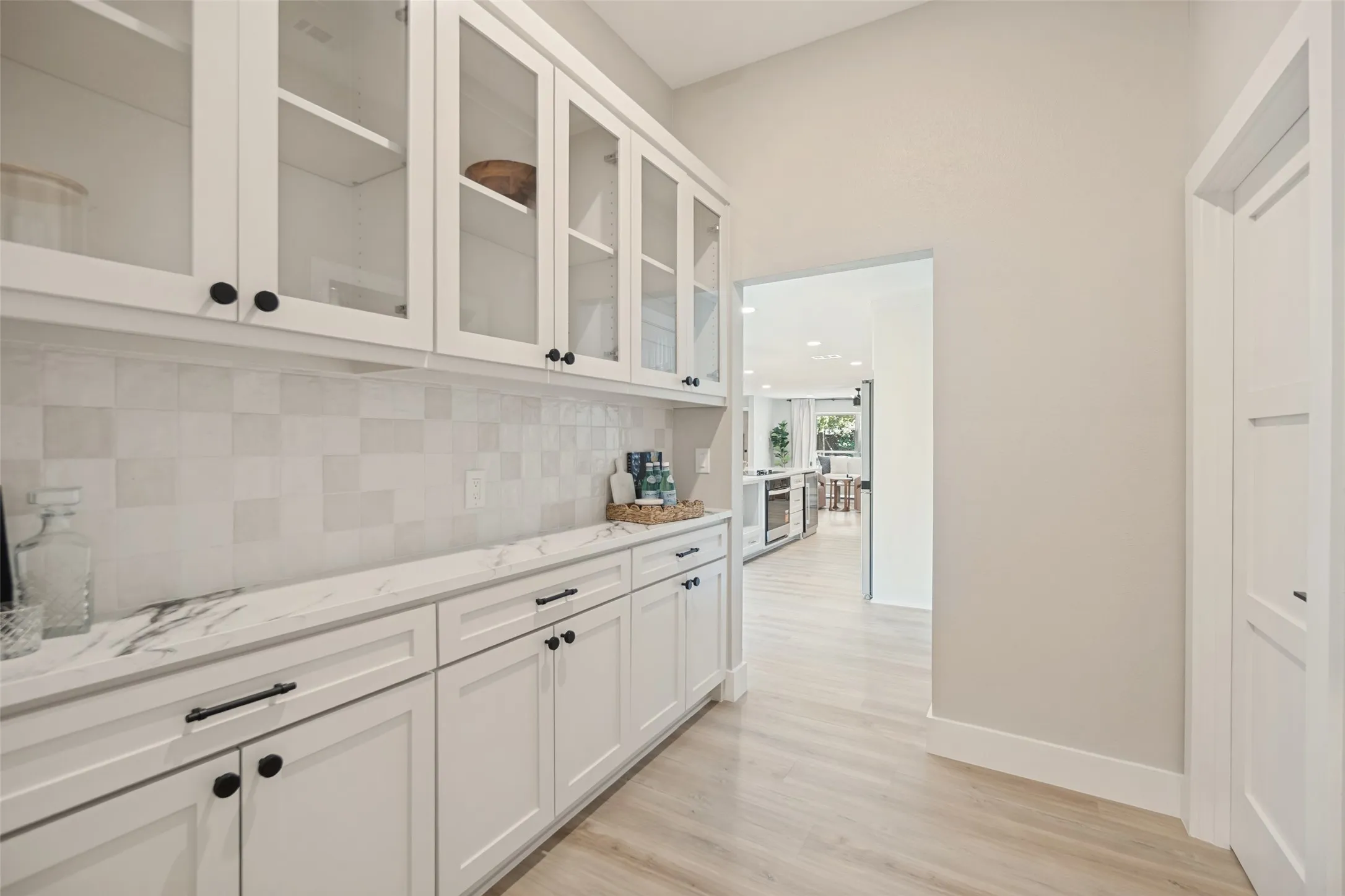 White shaker cabinetry with black hardware, glass-front upper cabinets, white tile backsplash, light wood-finish flooring, and a white countertop with gray veining