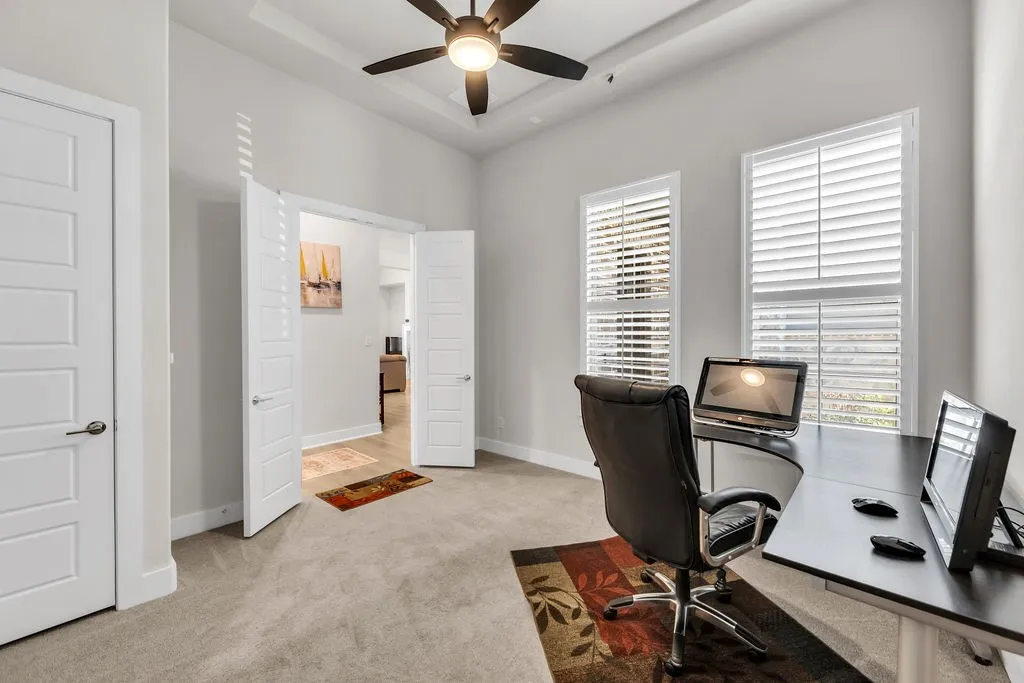 Tray ceiling with a ceiling fan, two windows with plantation shutters, light-colored carpeting, and a double-door entryway