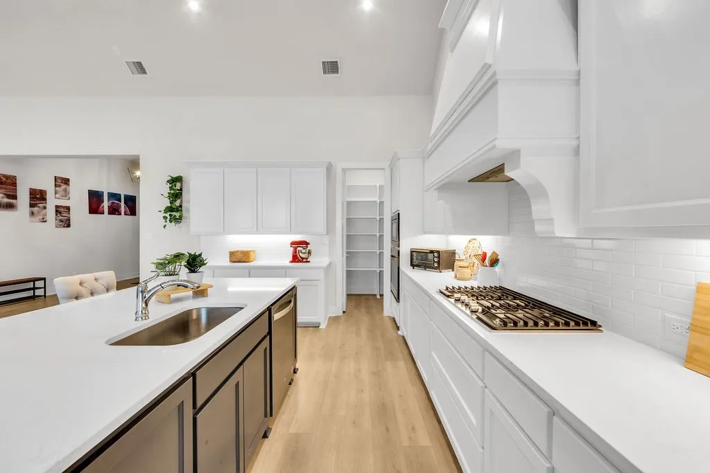Spacious kitchen featuring an expansive island with an undermount sink, white shaker cabinetry, light wood-finish flooring, and a gas range with a decorative range hood