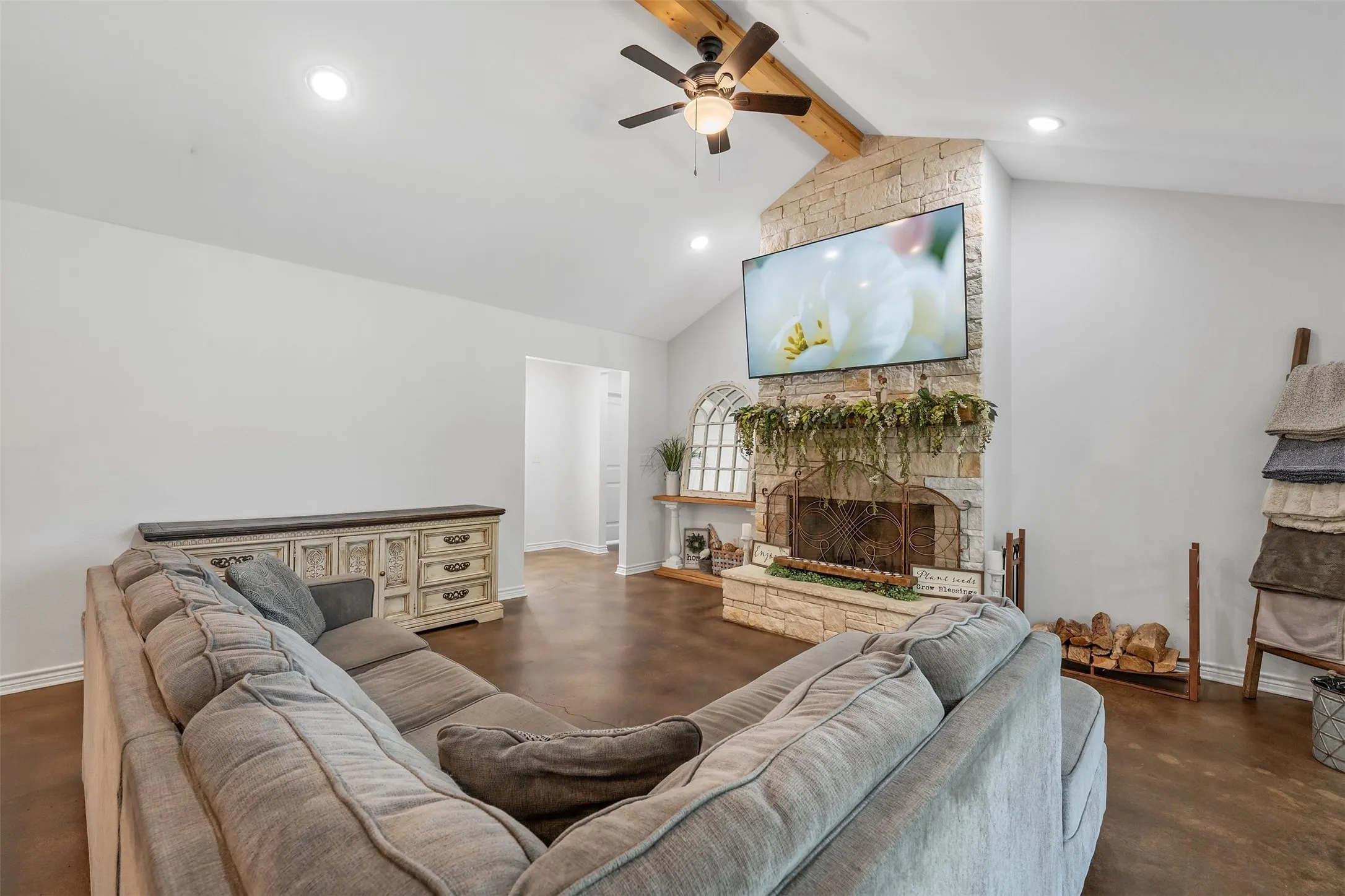 Vaulted ceiling with exposed wood beam, stone fireplace with a hearth, integrated ceiling fan, recessed lighting, and wood-finish flooring