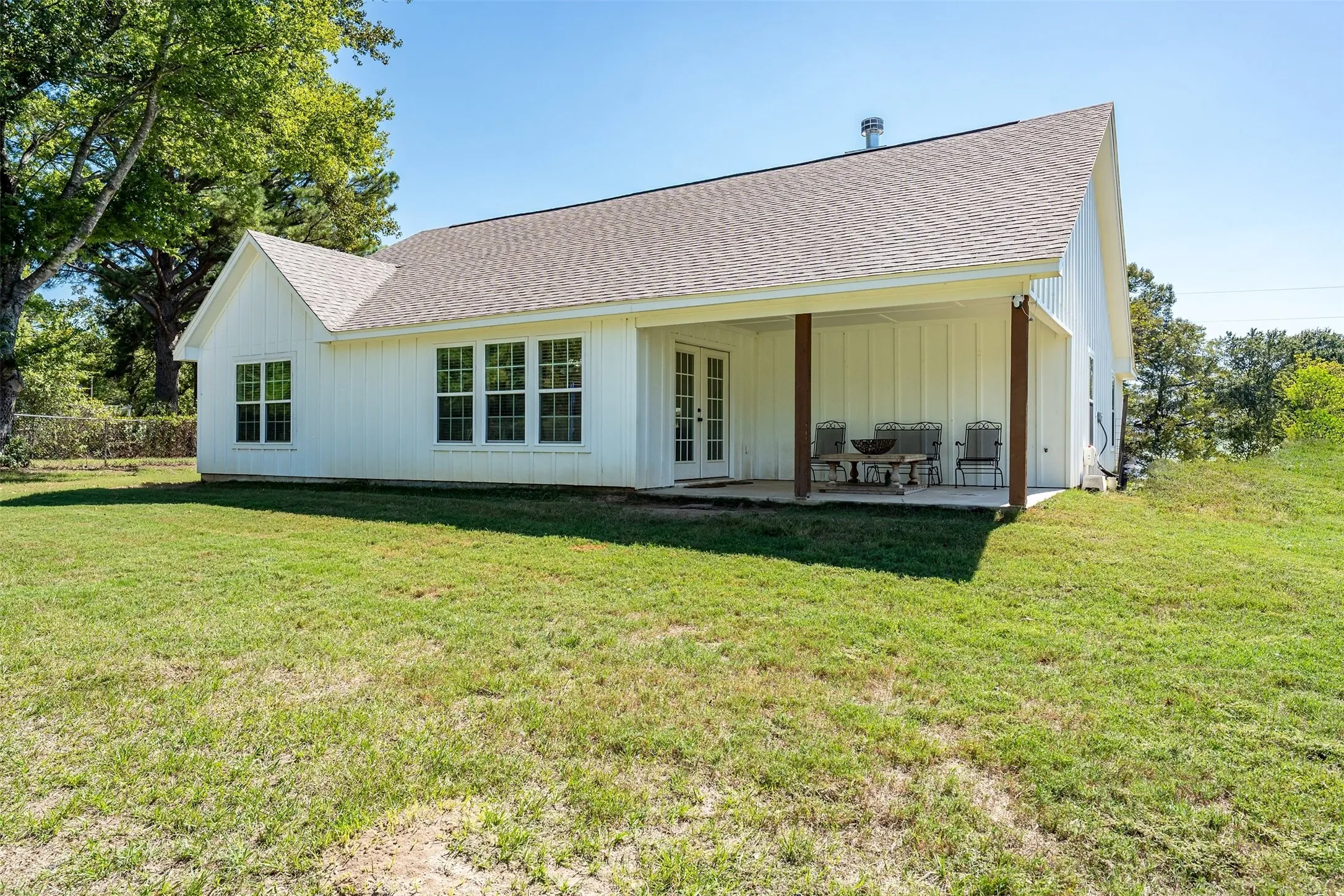 White board and batten siding, multiple pane windows, double French doors, covered patio with wood support beams, and a gabled roof