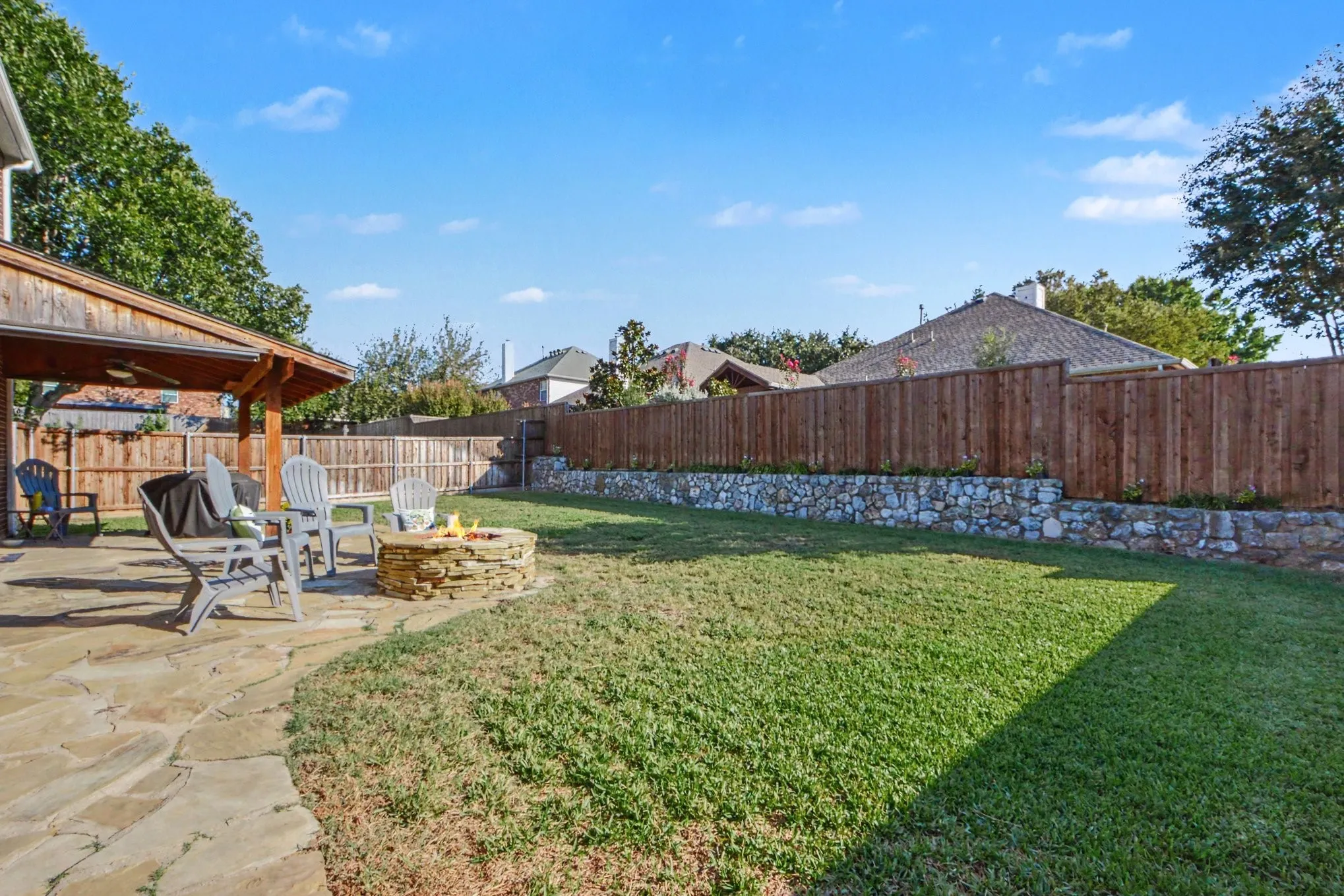 Expansive backyard featuring a stone patio, wooden pergola, built-in stone fire pit, and wood privacy fencing with a stacked stone retaining wall