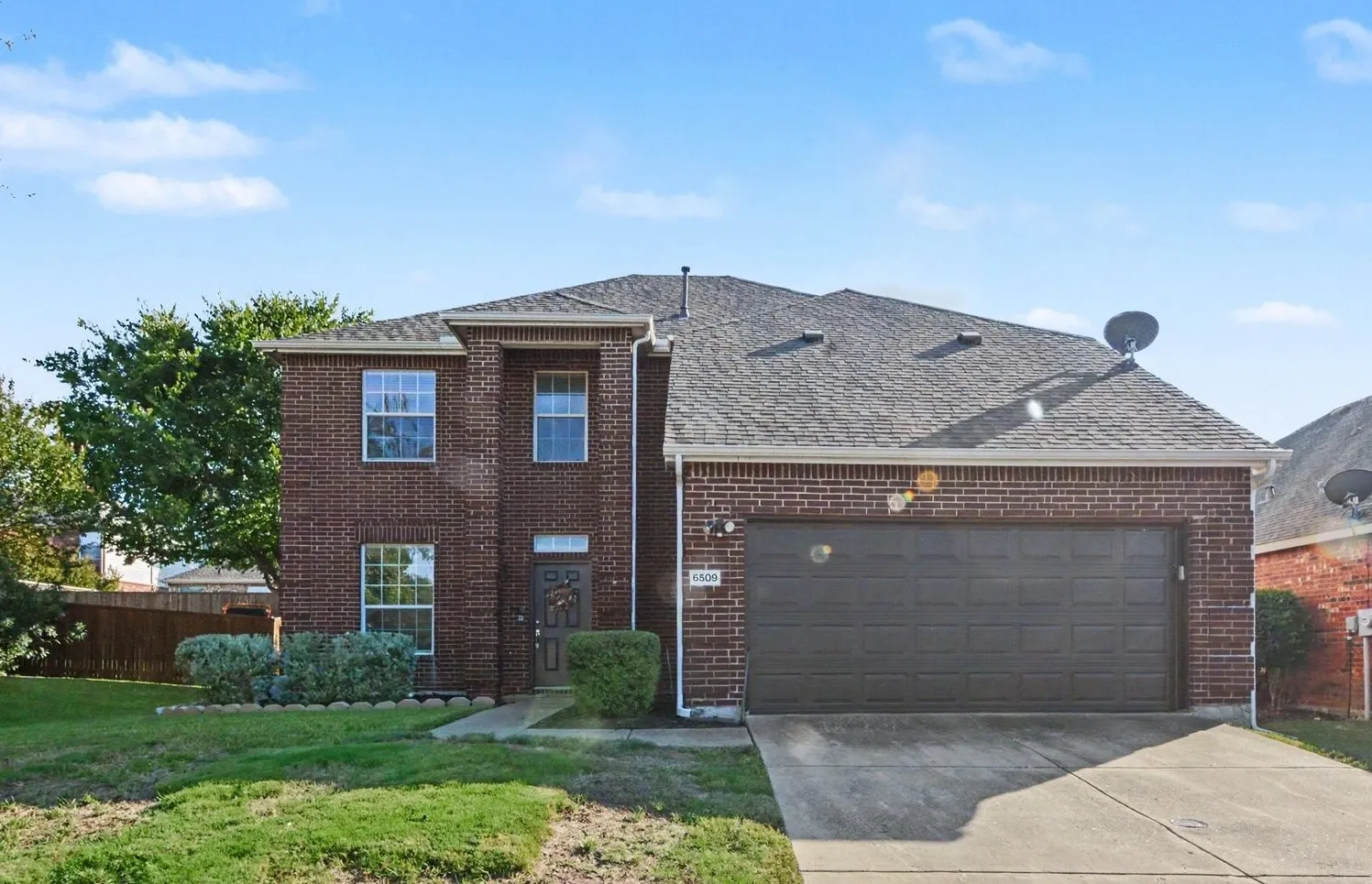 Brick exterior facade, dark shingle roof, attached garage with panel door, concrete driveway, and established front landscaping