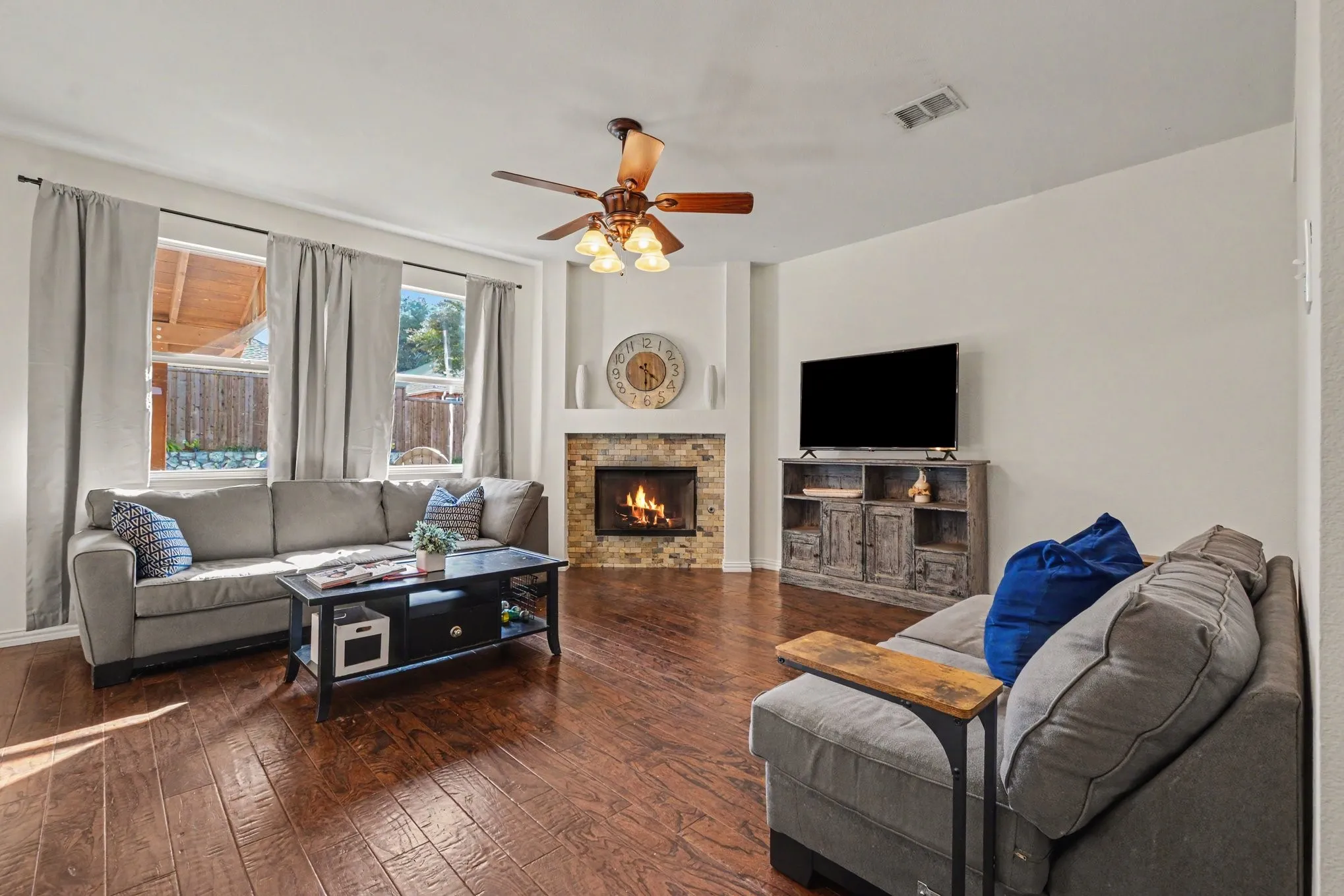 Corner fireplace with stone surround, wood-finish flooring, ceiling fan, and multiple windows