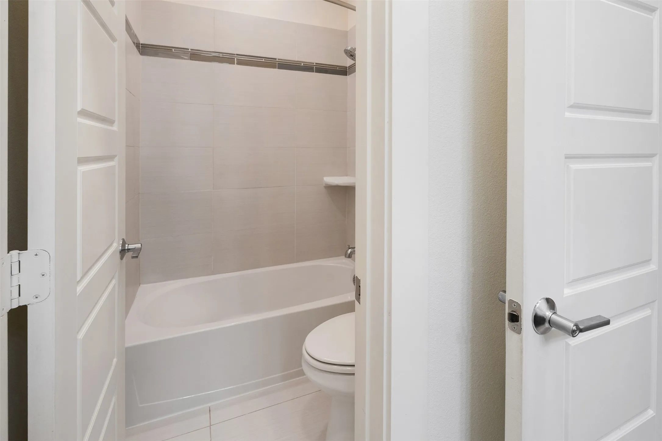 Full bathroom with a white bathtub, light-colored wall tiles, a decorative tile accent band, and a built-in shower shelf