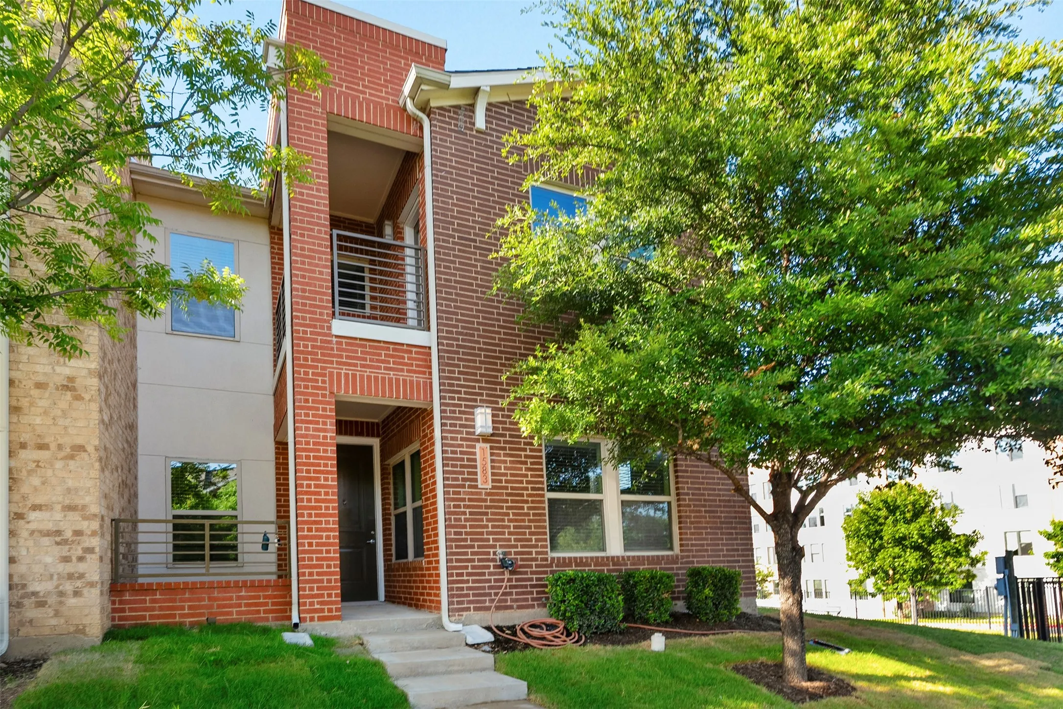 Contemporary facade featuring red brick and stucco, a prominent front entry, upper-level balcony with metal railing, and a ground-level patio with matching railing