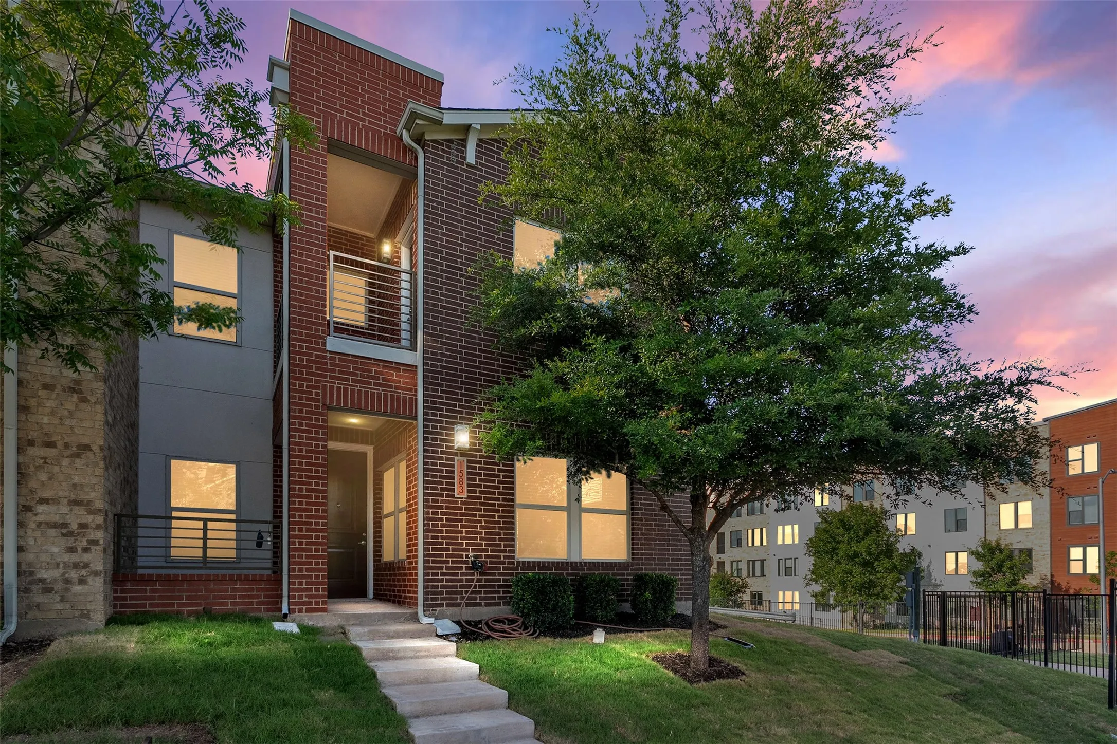 Brick and panel facade, concrete walkway, ground-level entry, second-story balcony with metal railing, and mature tree