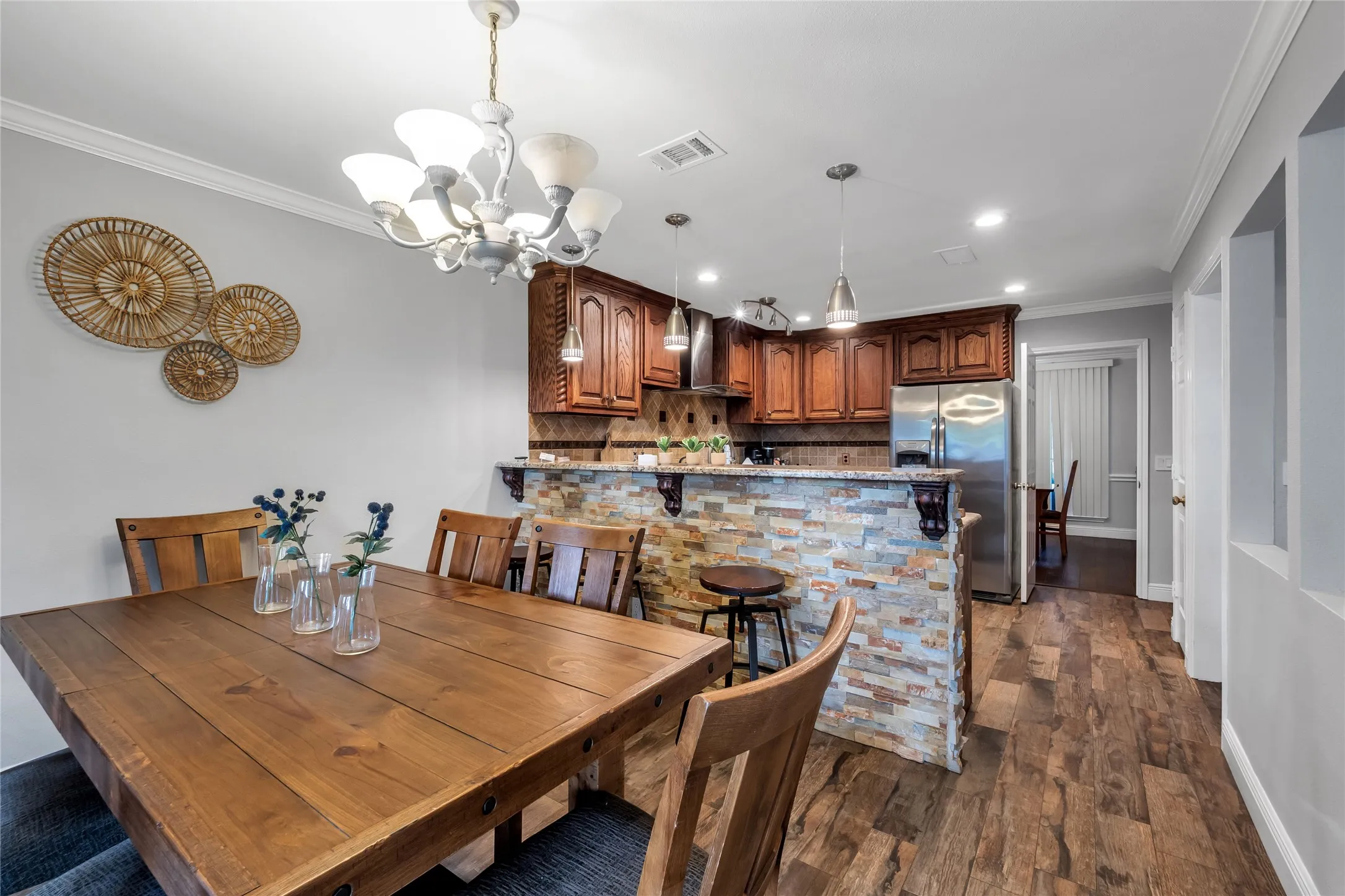 Open-concept dining and kitchen area featuring warm wood cabinetry, stainless steel appliances, a multi-colored brick backsplash, wood-finish flooring throughout, and a decorative chandelier overhead
