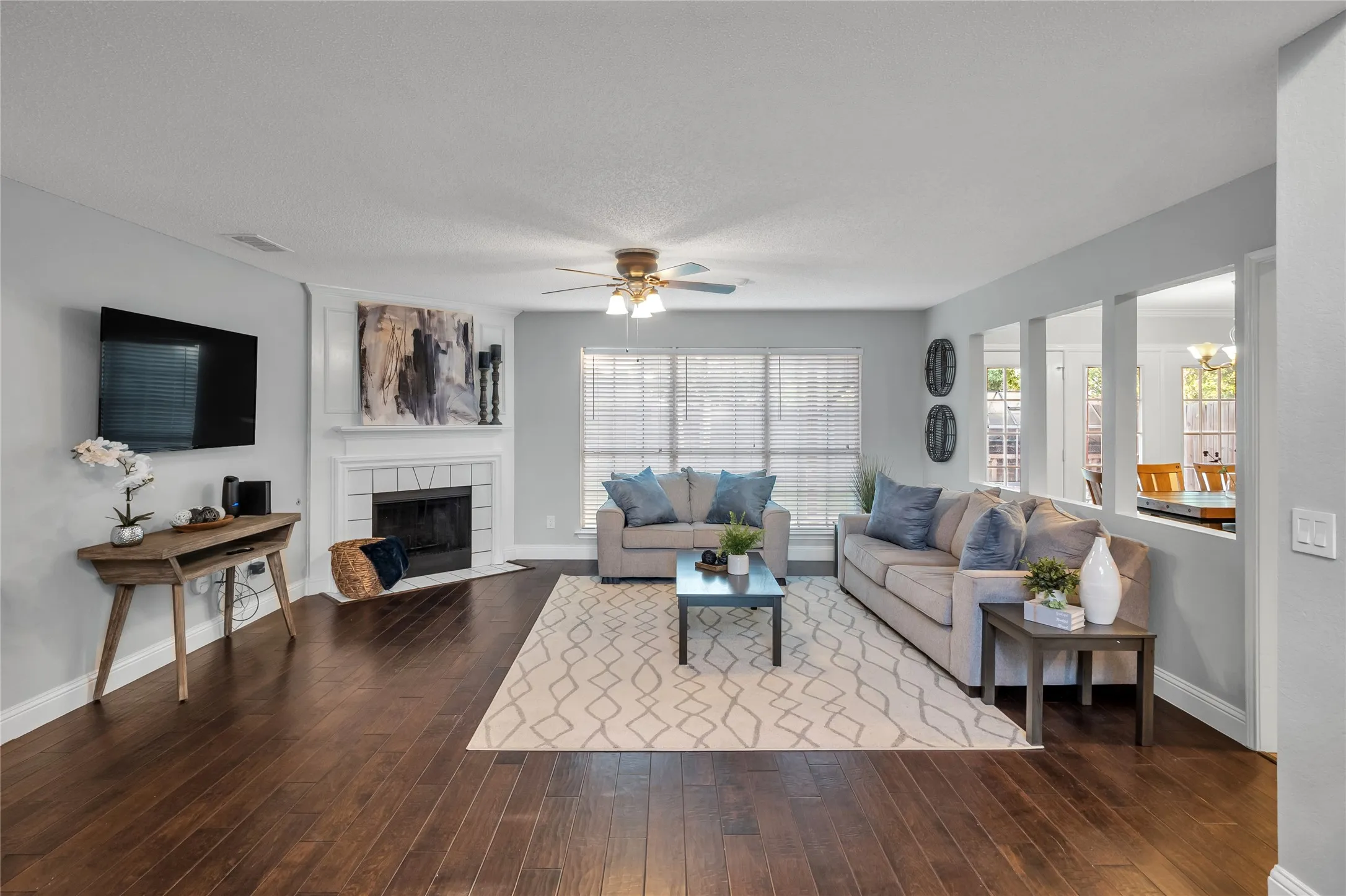 Dark wood-finish flooring, white fireplace, multiple windows with blinds, ceiling fan, and a pass-through opening to an adjacent room