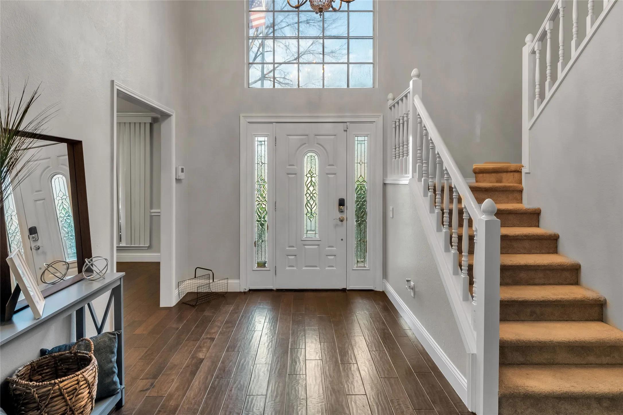 Bright two-story foyer featuring a transom window, hardwood flooring, white-painted staircase with wooden treads, and a glass-paneled front door with sidelights
