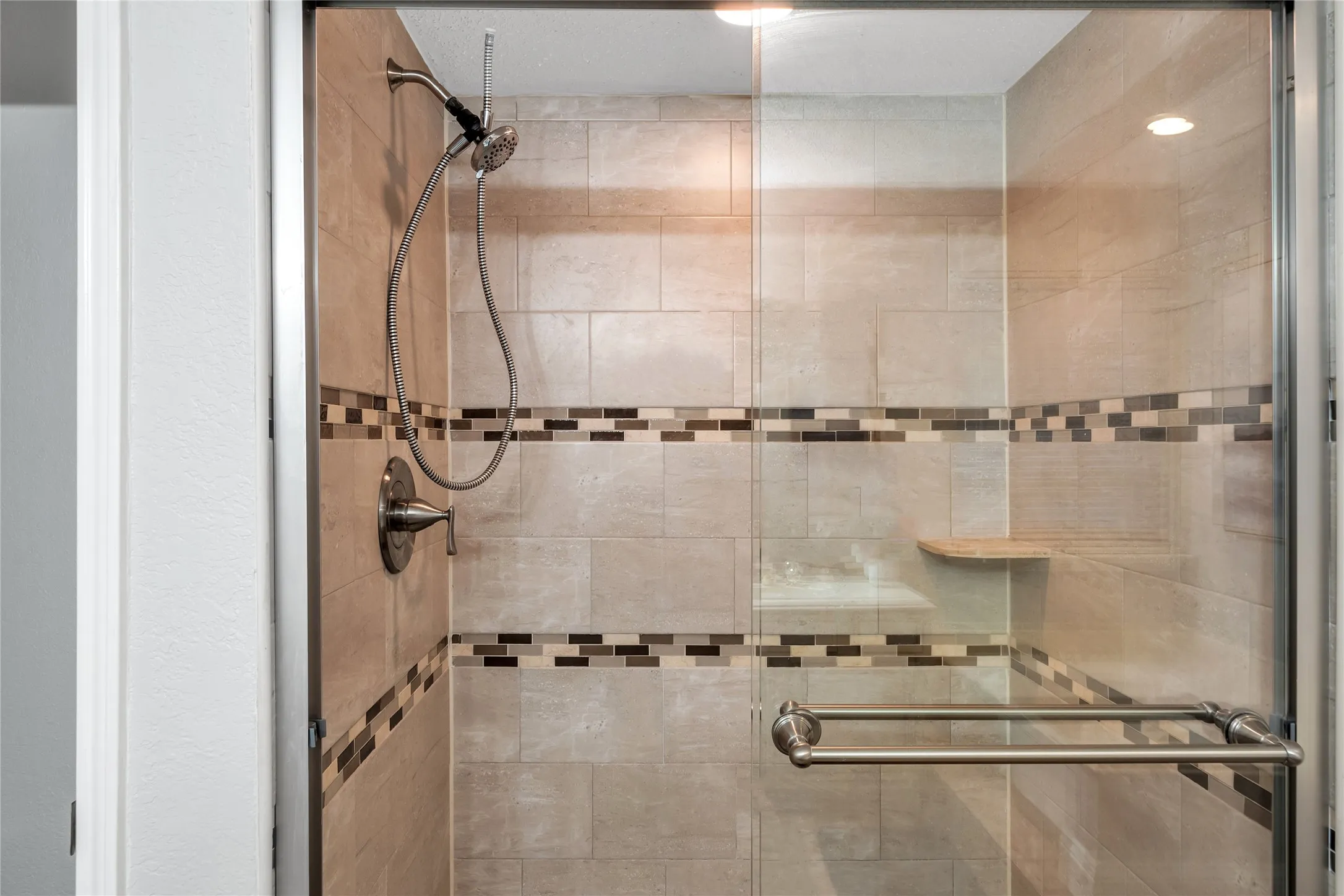 Tiled shower enclosure featuring a mosaic accent band, brushed nickel fixtures, glass shower door, and an integrated corner bench